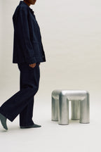 Person wearing a dark outfit standing next to a metallic stool on a plain background