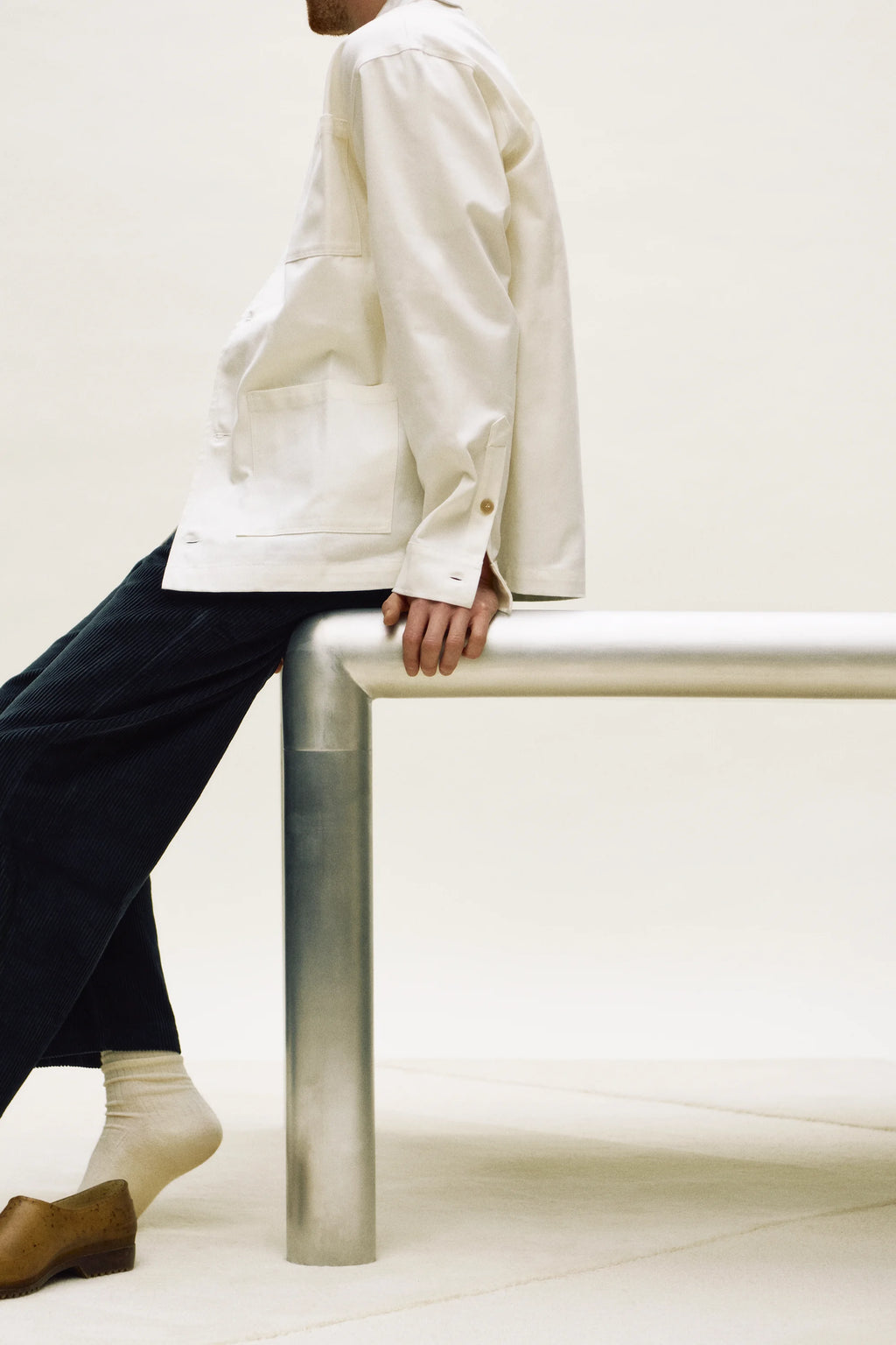 Person wearing a white jacket and dark pants sitting on a white bench against a white background