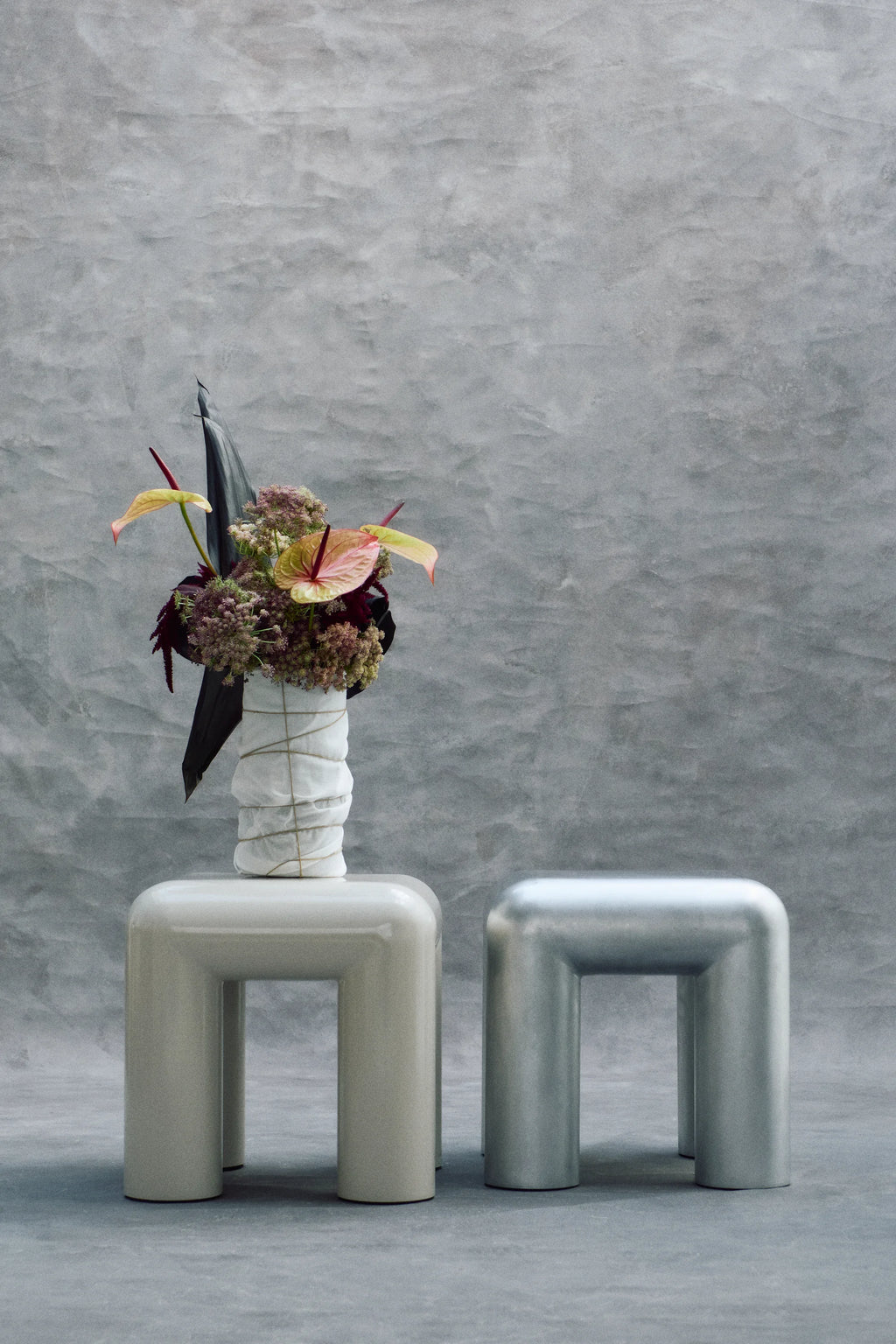 Two modern stools with a vase of flowers against a gray concrete wall.