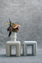 Two modern stools with a vase of flowers against a gray concrete wall.