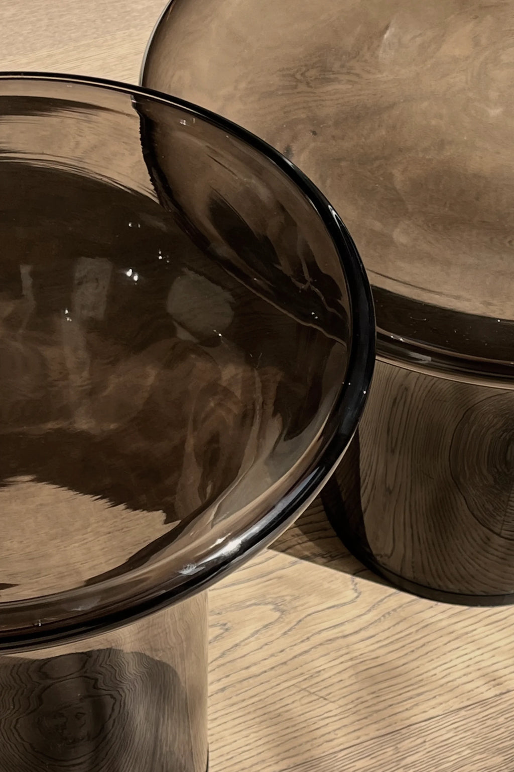 101 CPH Trumpet Side Table - Glass. Close-up view of two contemporary dark brown translucent glass tables on a light wood floor, reflecting the wood grain. This luxury accent furniture offers versatile styling.