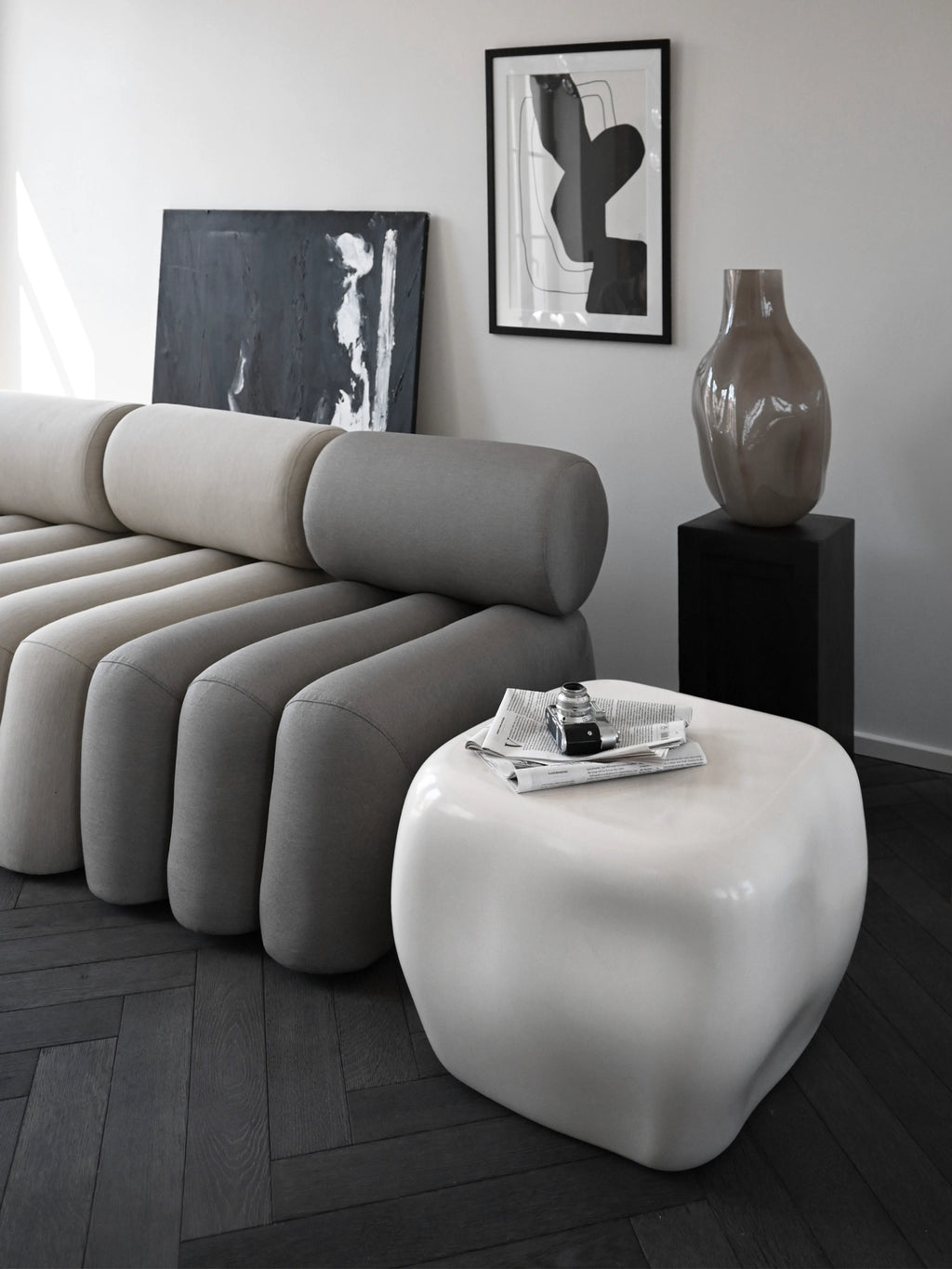 101 CPH Ishi Coffee Table - Ceramic. Organic-shaped, glossy white handcrafted ceramic coffee table with a newspaper and vintage camera on top, next to a grey modular sofa. A luxury centerpiece for modern living rooms.