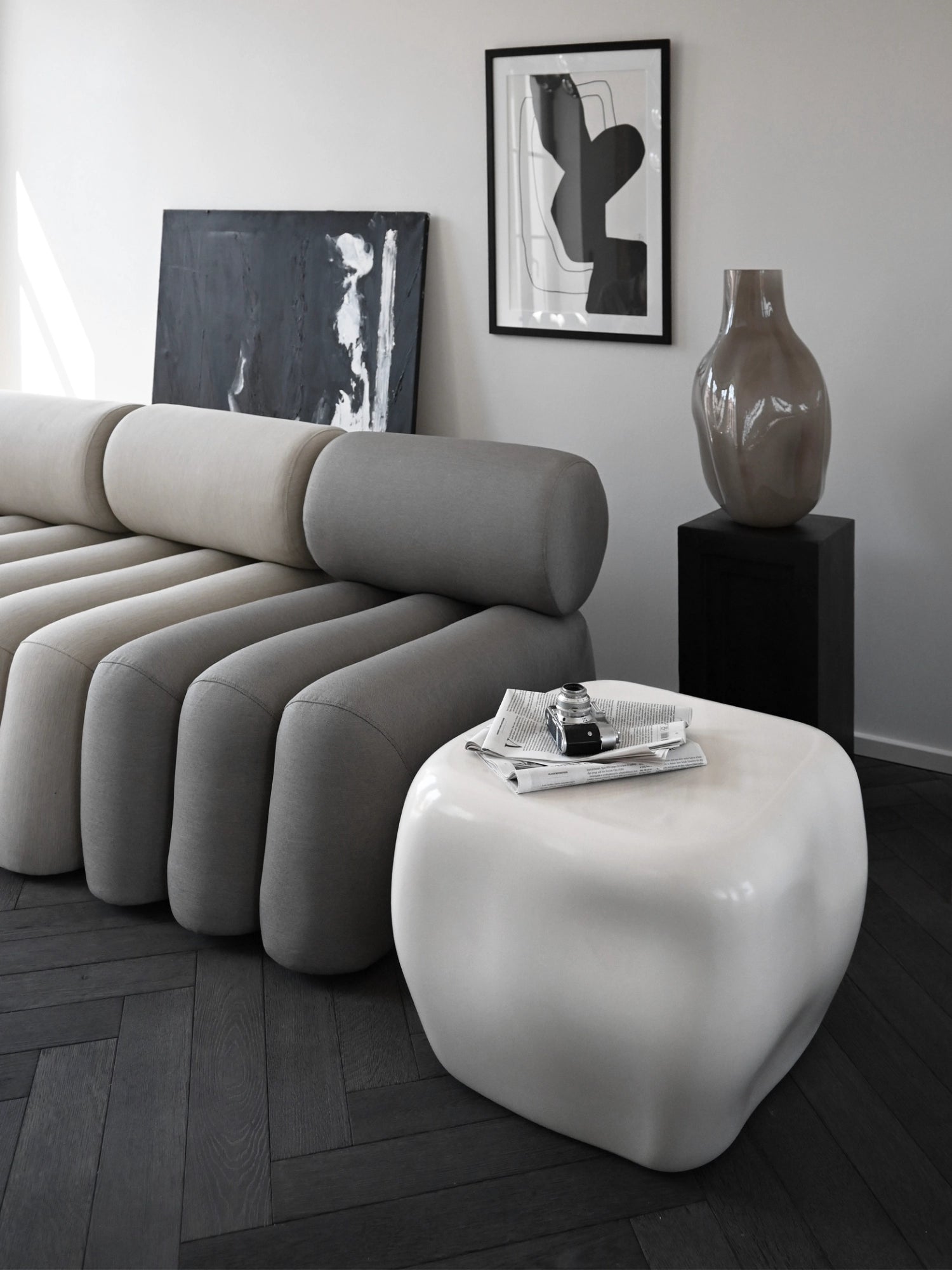 101 CPH Ishi Coffee Table - Ceramic. Organic-shaped, glossy white handcrafted ceramic coffee table with a newspaper and vintage camera on top, next to a grey modular sofa. A luxury centerpiece for modern living rooms.