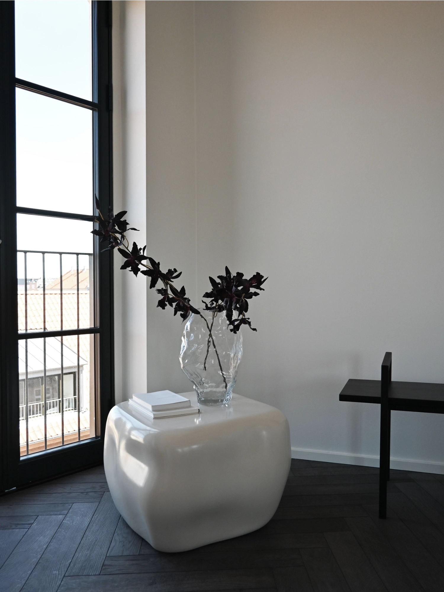 101 CPH Ishi Coffee Table - Ceramic. Lifestyle shot of a white, organic-shaped, glossy ceramic coffee table with a clear vase and dark foliage, next to a black-framed window. A luxury, contemporary statement piece.