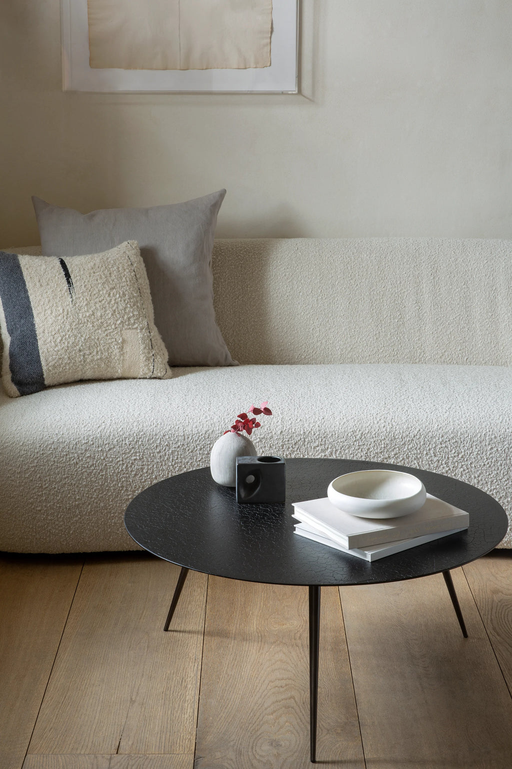 Ethnicraft Luna Coffee Table, a round, elegant black table with a textured, lava-like finish. It features slender, tapered black metal legs, styled in a contemporary living room setting with a cream sofa and decorative pillows.