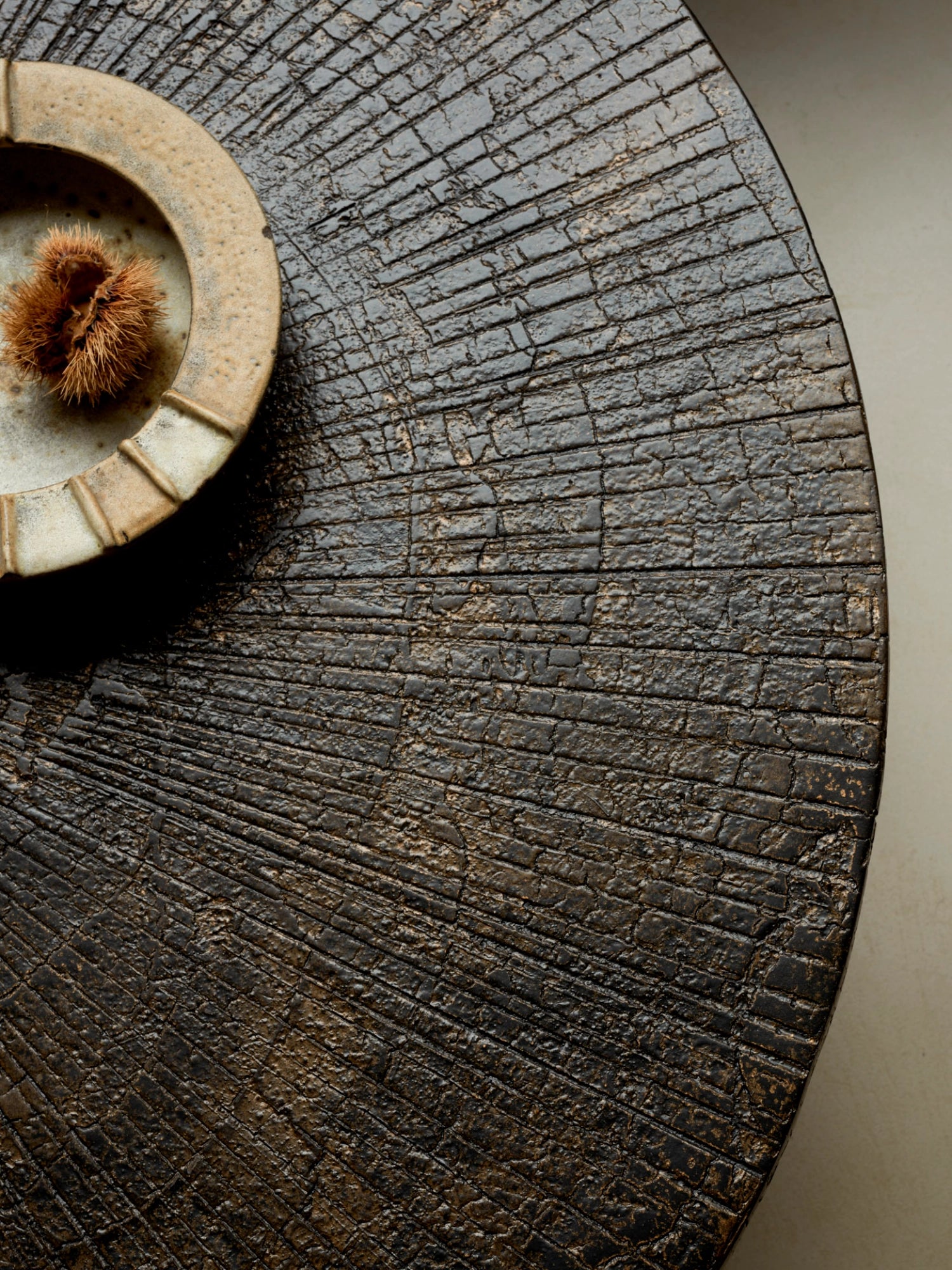 Ethnicraft Slice Coffee Table, a close-up of its round, dark wood surface with a unique, deeply textured, cracked-earth pattern. A rustic ceramic bowl holding chestnuts rests on its left side, emphasizing its organic, artisanal appeal.