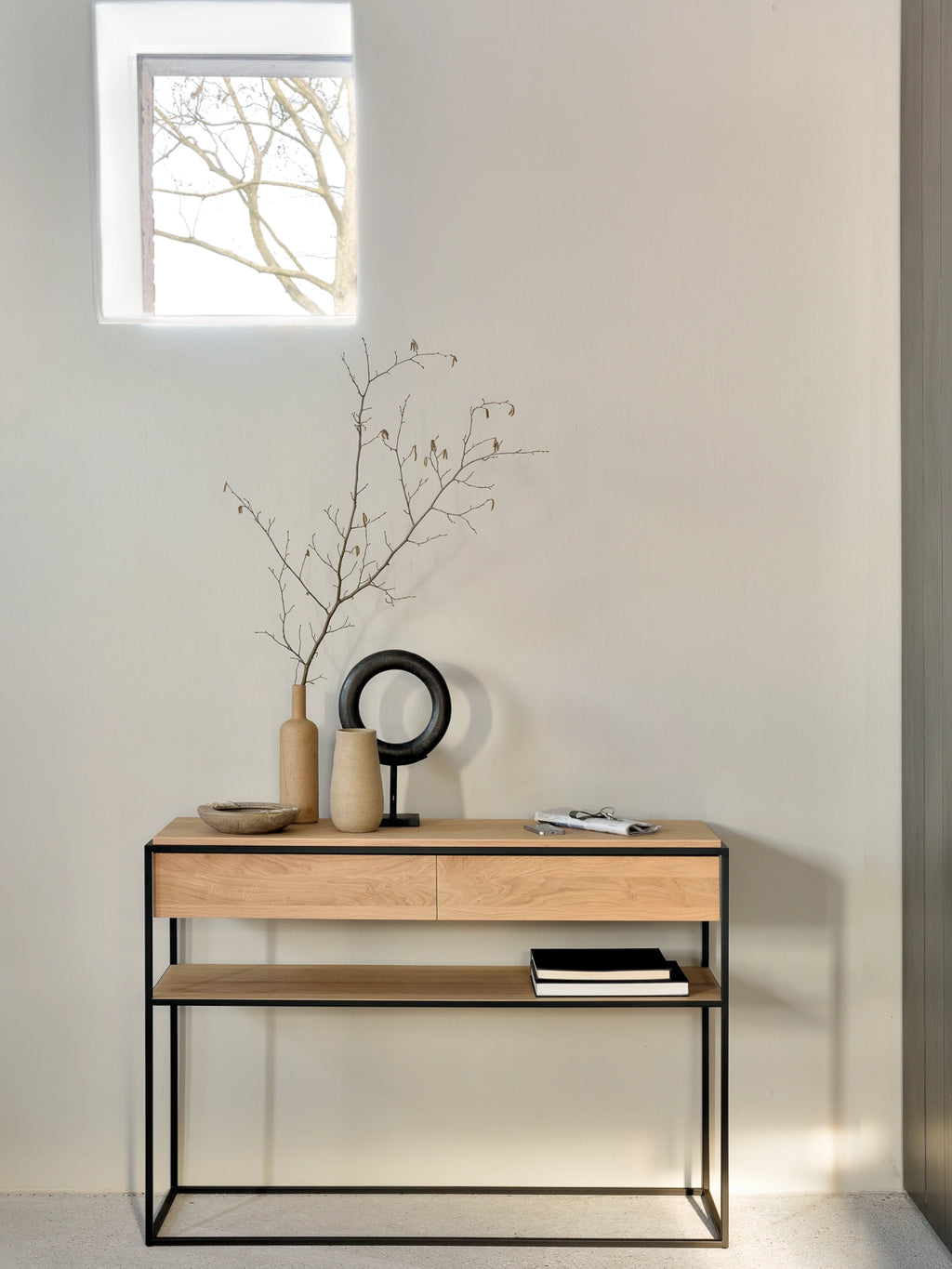 Ethnicraft Monolit Console, a modern minimalist console table crafted from solid light oak wood with a natural finish, featuring two seamless, handleless drawers and an open lower shelf, all supported by a clean-lined matte black metal frame. Its contemporary industrial chic design makes it a versatile entryway console table or sophisticated accent for living rooms and hallways.