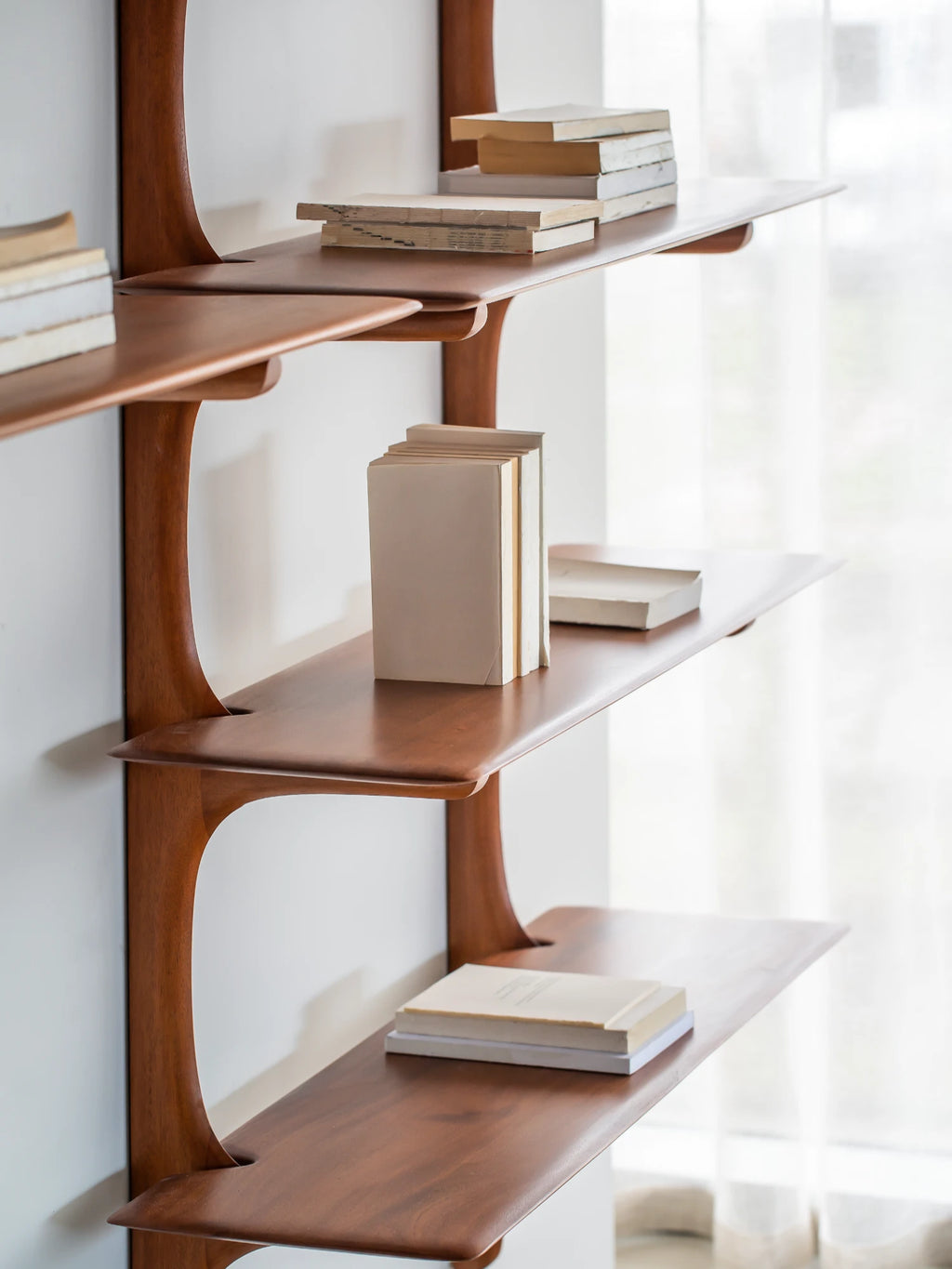 Ethnicraft PI Wall Shelf, a modular wall shelving system crafted from warm solid teak wood, featuring elegantly curved, organic support structures and smooth, rectangular shelves. Its fluid lines and rich reddish-brown finish embody an organic modern and mid-century aesthetic. Perfect for stylish display and storage in contemporary living rooms, home offices, or minimalist bedrooms.