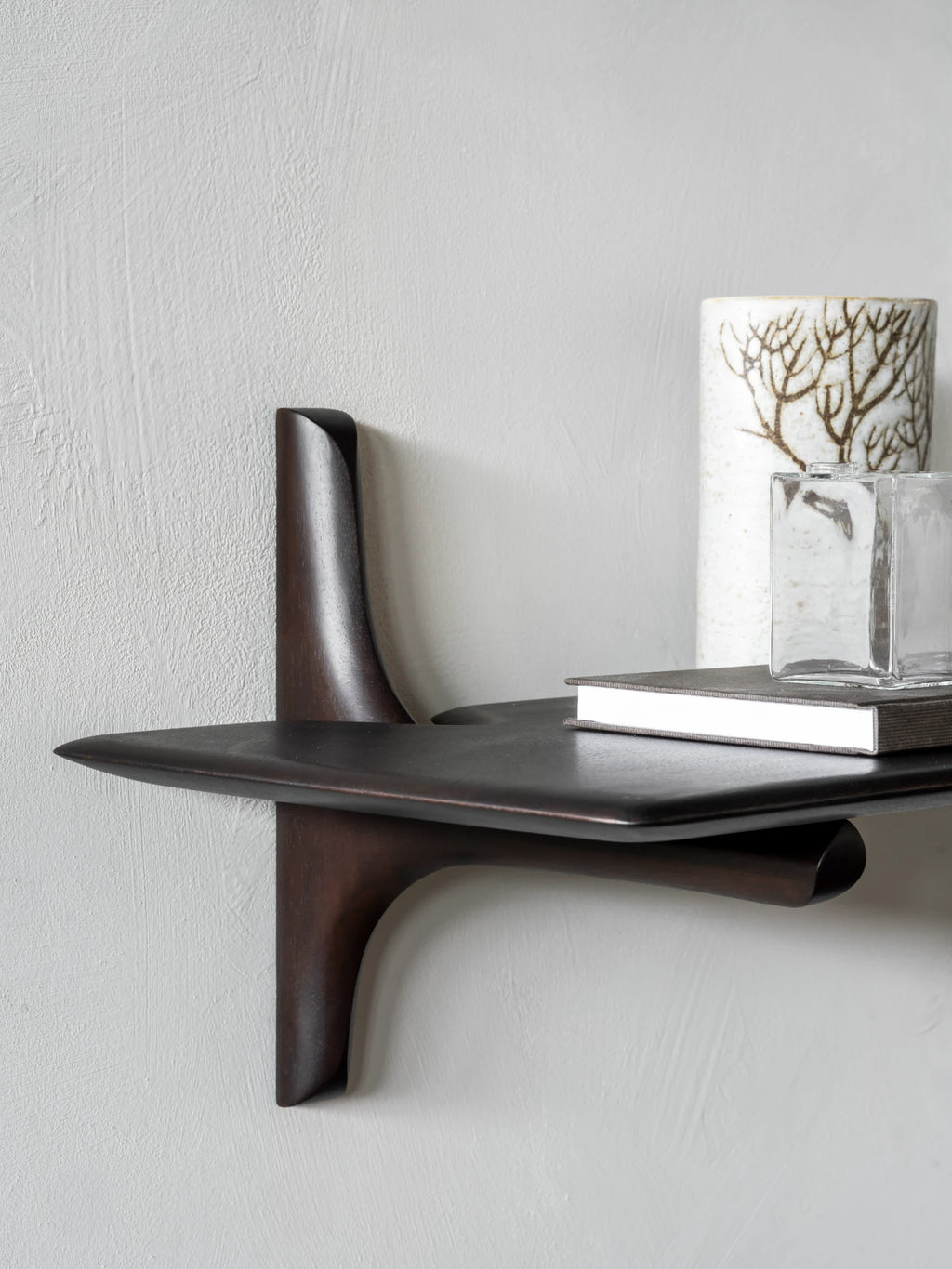 Ethnicraft PI Wall Shelf, a dark brown solid wood wall shelf featuring a sculptural organic modern design with gracefully curved, flowing forms that create a unique floating aesthetic. This contemporary wall shelf is crafted from rich, smooth dark wood, highlighting its distinctive silhouette. Perfect for minimalist interiors, modern living rooms, or as a striking entryway accent.
