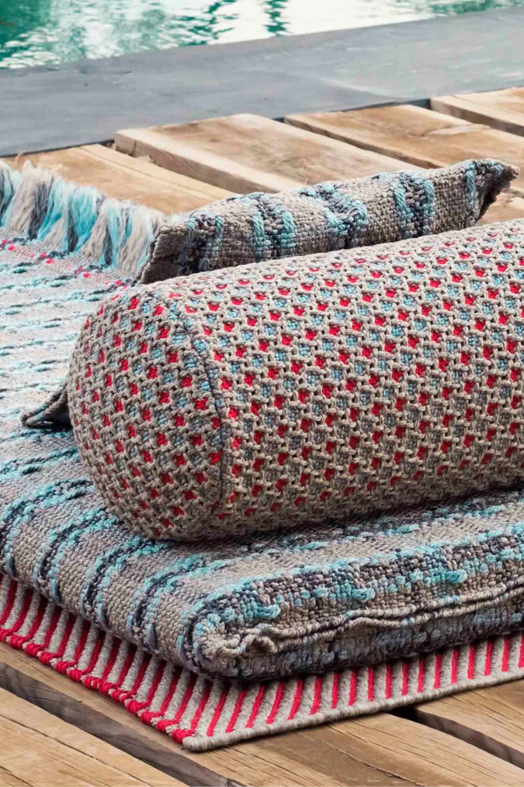 GAN Rugs Garden Layers Composition 05, a textured hand-woven outdoor bolster cushion featuring a grey base intricately patterned with coral red and aqua blue woven dots.