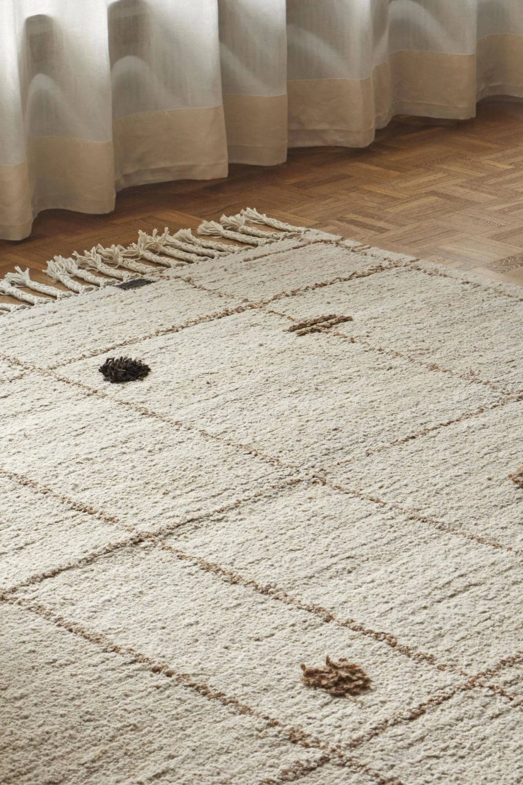 Sera Helsinki Virta Rug, a luxurious hand-knotted natural wool area rug featuring a plush, shaggy cream pile with intricate brown geometric grid patterns and dark brown tufted accent dots. This organic modern rug showcases a rustic texture and natural fringe detailing on one end. Ideal as a cozy boho rug for living rooms, Scandinavian bedrooms, or minimalist spaces seeking warmth and texture.