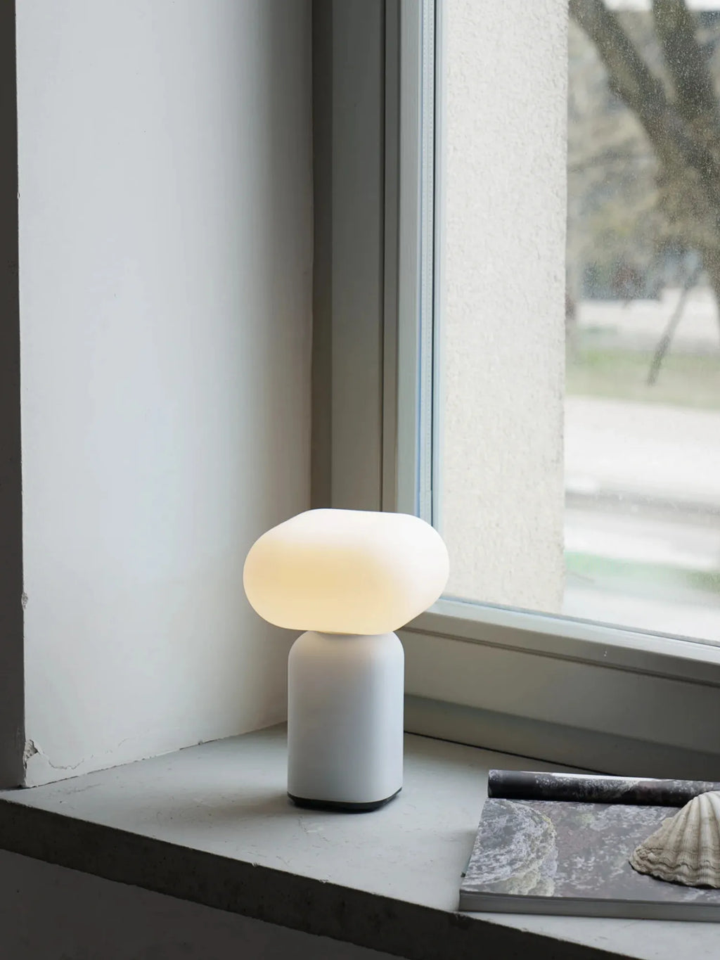 TAMO Velis Portable Lamp, a minimalist white cylindrical base with an illuminated warm white amorphous glass diffuser, displayed on a windowsill with a magazine. A premium, modern lighting solution for any space.