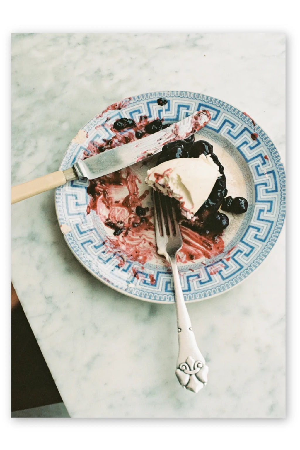 The Poster Club Weekend Pancakes, a charming still life photography poster capturing a delicious blueberry dessert with cream, served on a vintage blue and white Greek key patterned plate, alongside silver cutlery on a light marble table. This rustic food photography print adds cozy charm to kitchen, dining room, or breakfast nook decor.
