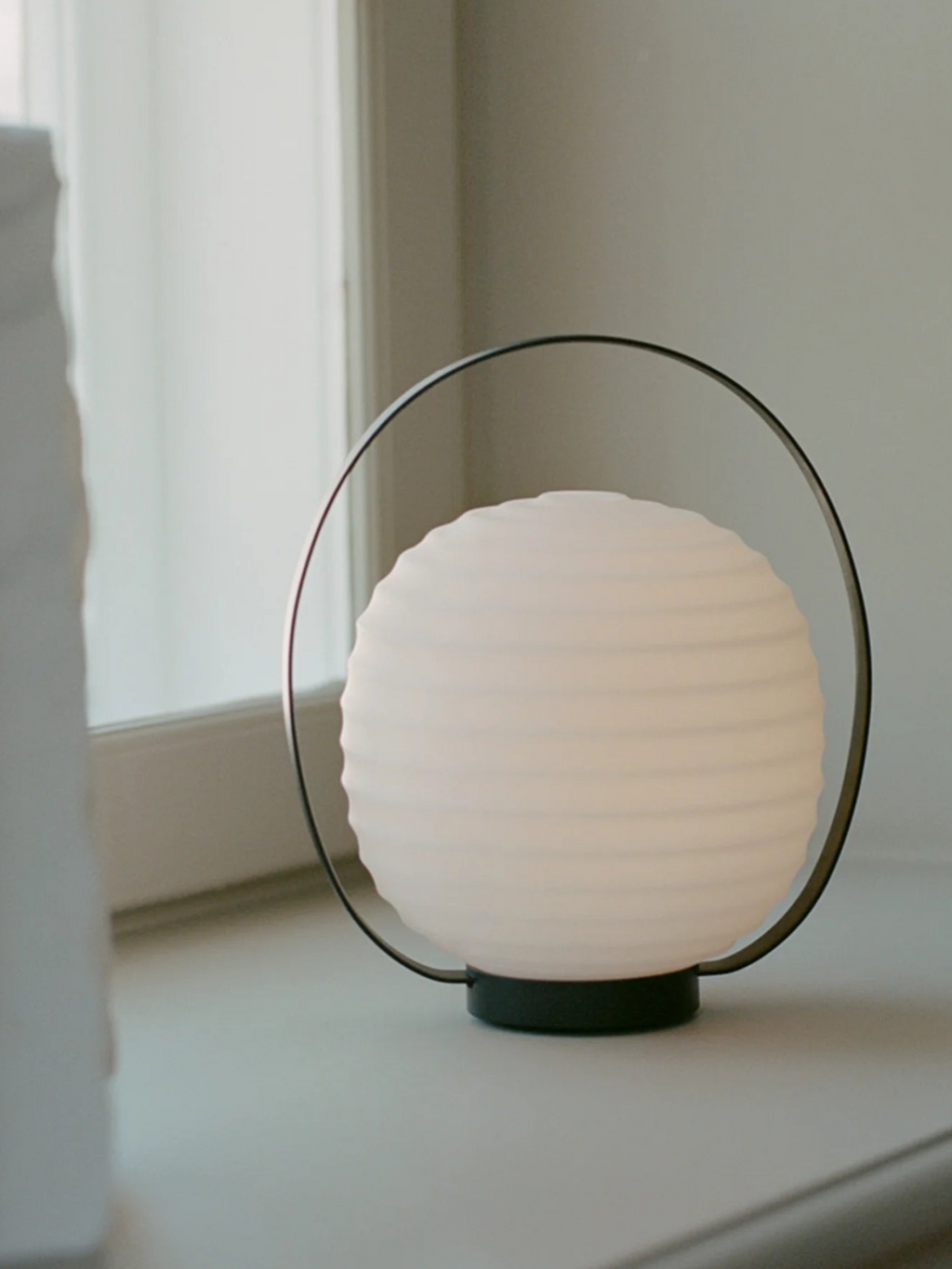 New Works Lantern Portable Lamp, a luxurious modern lamp with an off-white ribbed spherical diffuser and sleek black metal handle, shown on a light-colored window sill. A premium design for ambient lighting.