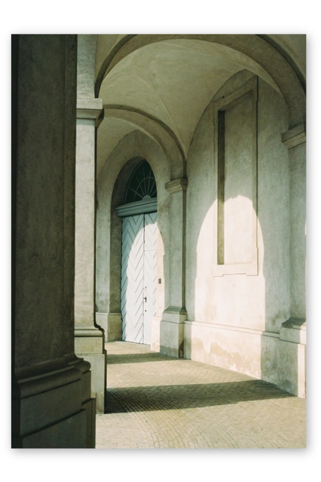 The Poster Club Morning Sun, an architectural photography print showcasing a serene classical European building corridor with textured cream-colored arched walls, robust stone columns, and a light blue diagonal patterned wooden door.