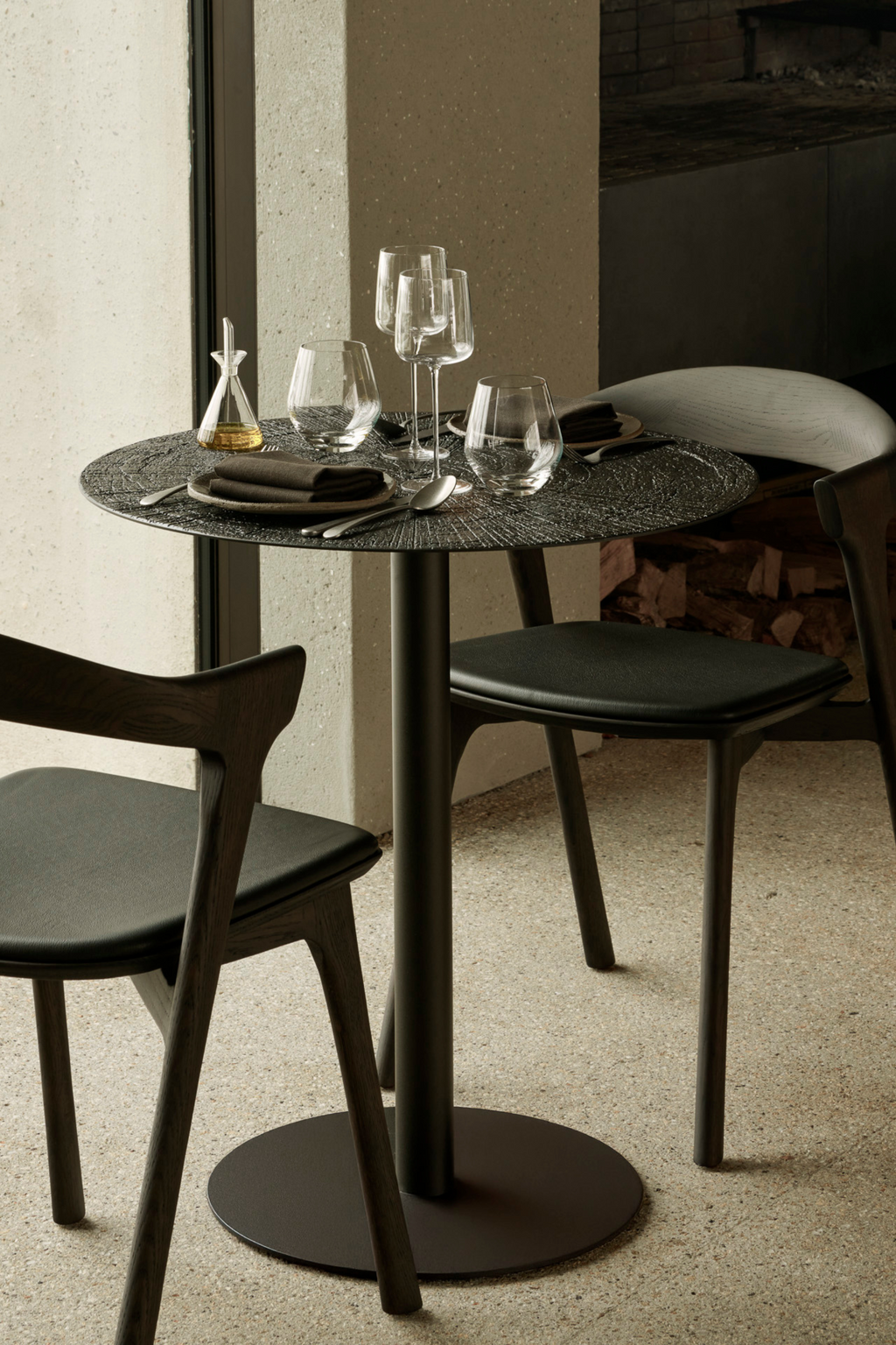 Ethnicraft Blend Round Dining Table, a contemporary industrial chic dining table featuring a unique dark charcoal textured metal tabletop with an organic, almost volcanic finish, supported by a sleek matte black pedestal base with a circular foot. This minimalist round dining table is perfect for modern dining rooms, stylish kitchens, or upscale restaurant settings.