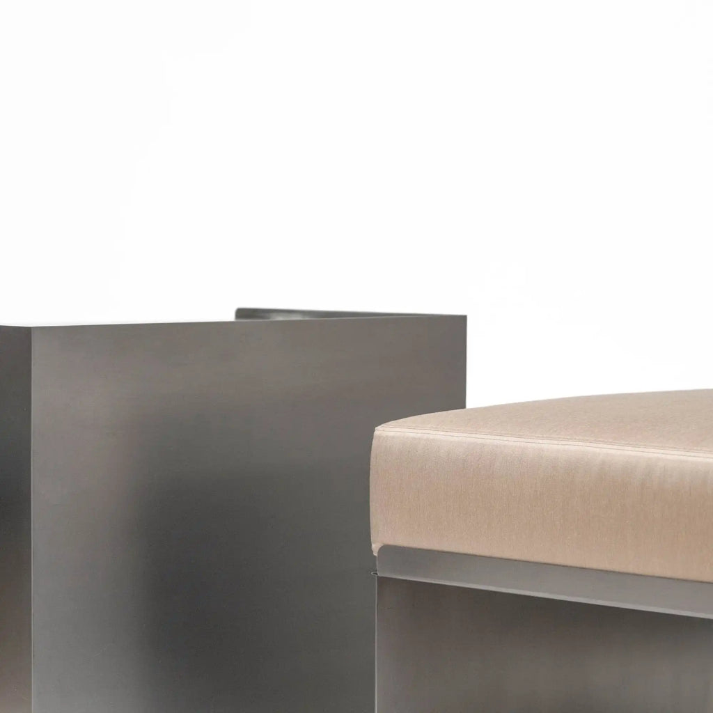 Around Works C.01 Lounge, a modern minimalist pouf featuring a robust matte dark grey metal base contrasted with a plush light beige fabric upholstered top and gently rounded edges. This contemporary ottoman offers versatile seating for a living room, bedroom accent, or stylish lounge area, blending industrial chic with organic modern comfort.
