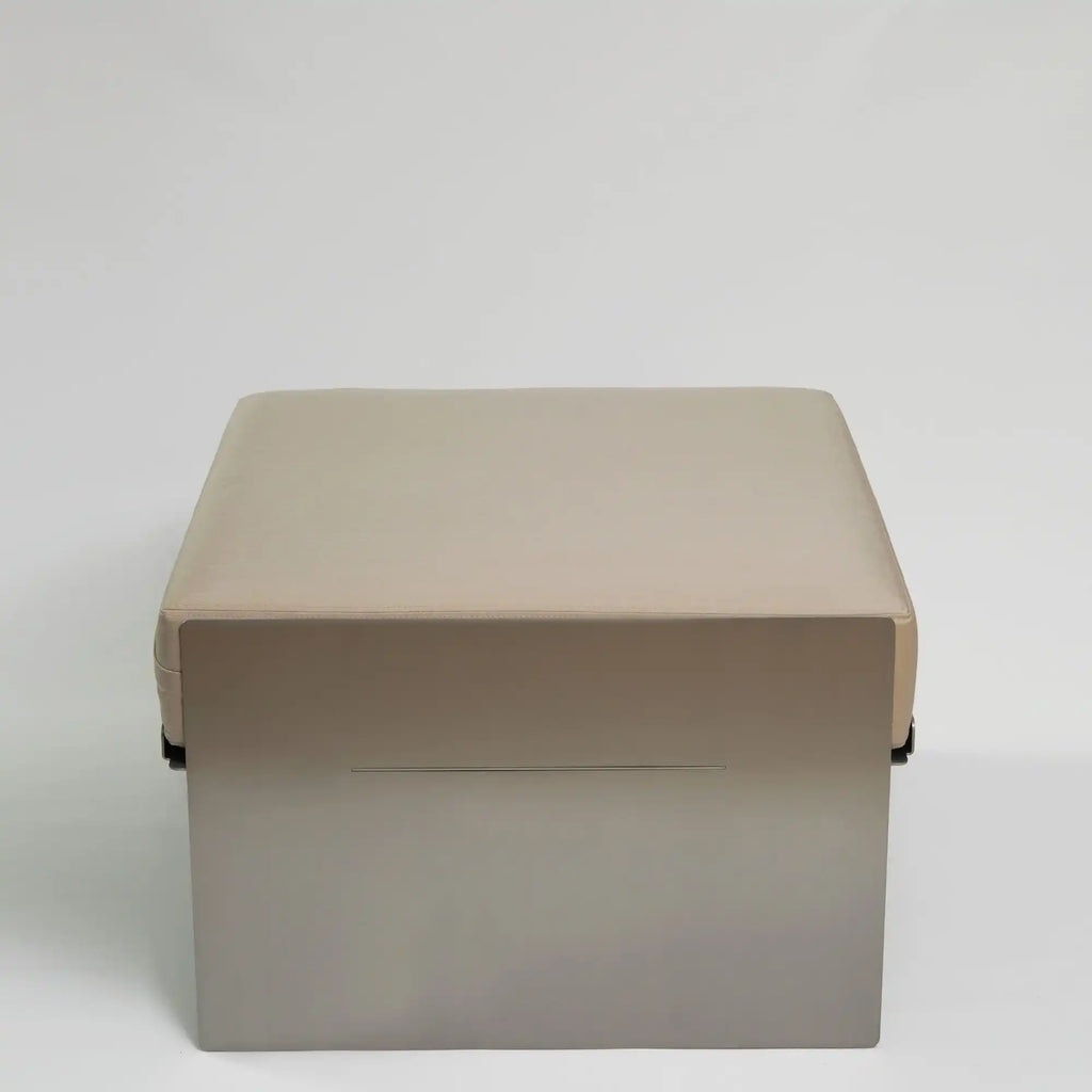 Around Works C.01 Lounge, a modern minimalist square pouf featuring a plush beige faux leather upholstered cushion atop a sleek matte taupe metal panel base with a subtle linear detail. This contemporary ottoman offers versatile accent seating and a clean aesthetic for living rooms, bedrooms, or lounge areas.