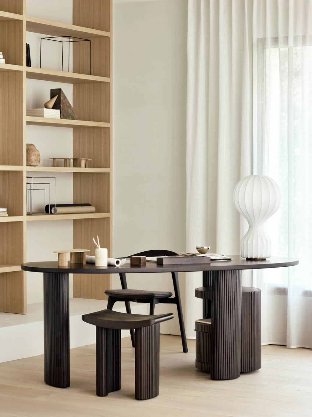 Ethnicraft Boomerang Desk in dark stained oak, featuring an elegant oval tabletop and fluted cylindrical legs, styled with a black chair and a unique large white lamp in a modern office.