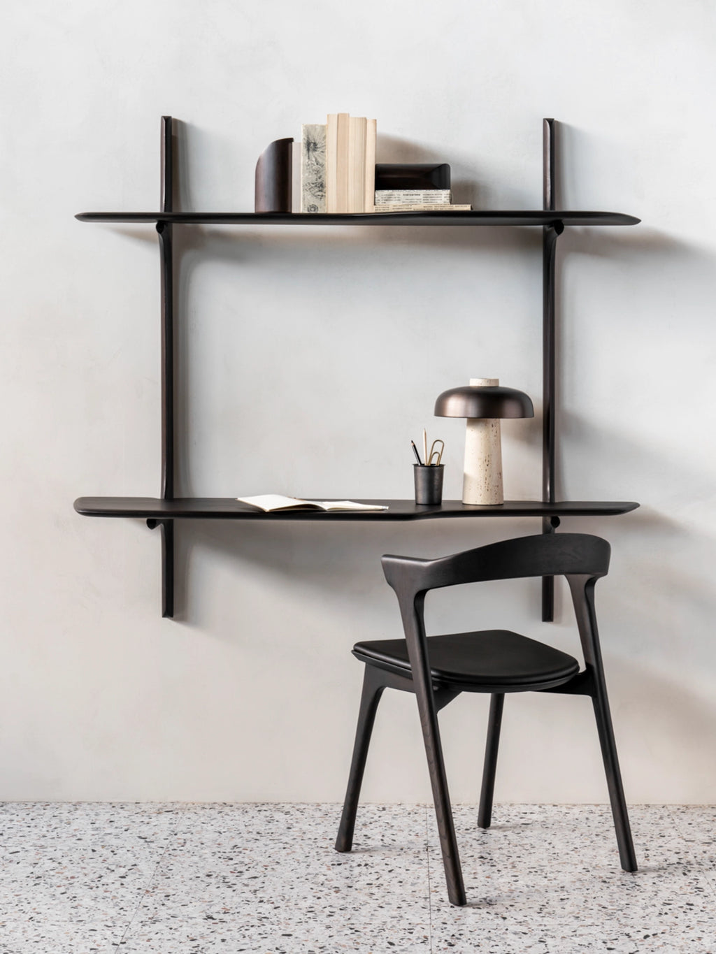 Ethnicraft PI Wall-Mounted Desk, a sophisticated dark brown varnished mahogany desk with two shelves, adorned with books and a stylish lamp, paired with a matching chair, against a textured white wall.