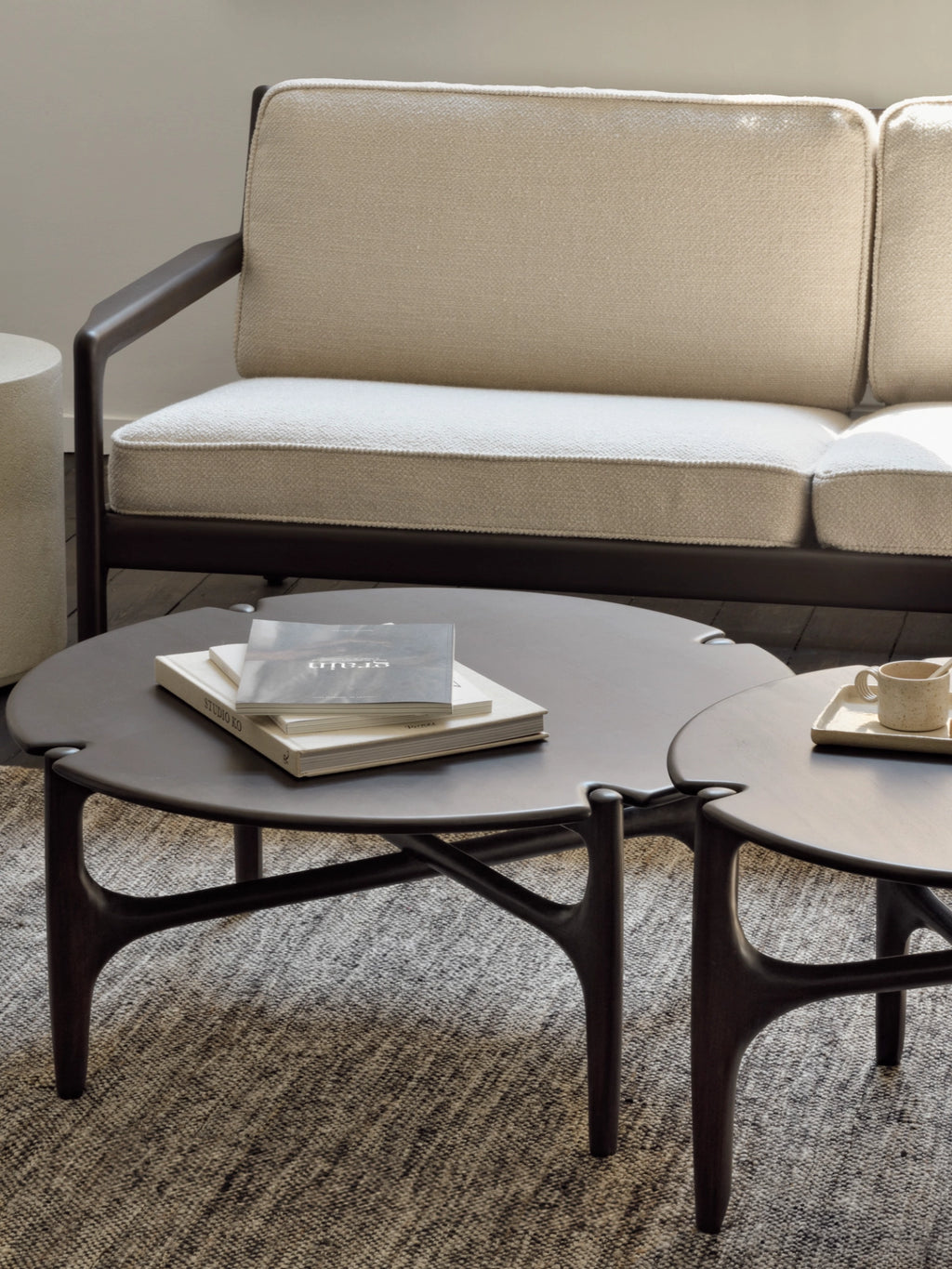 Ethnicraft PI Coffee Table, a pair of elegant dark brown round wooden tables with unique curved legs, displayed on a textured grey rug next to a cream sofa in a modern living room setting.