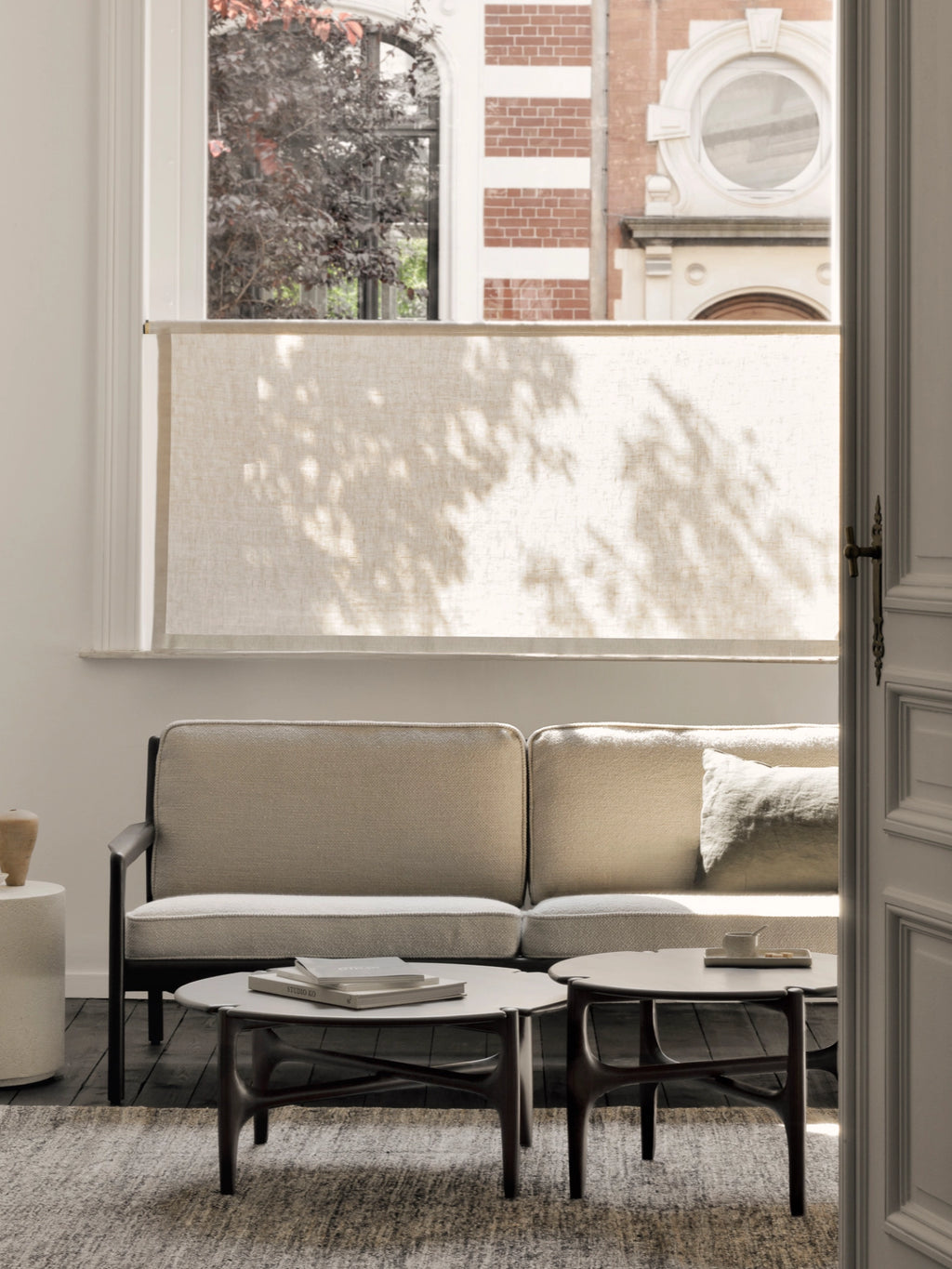 Ethnicraft PI Coffee Table, a set of two dark brown round wooden coffee tables with distinctive sculpted legs, positioned in front of a light-colored sofa in a sunlit room with a large window.