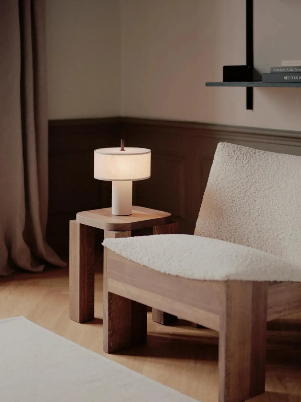 New Works Margin Portable Table Lamp, featuring a warm white cylindrical base and sheer fabric shade with a brown top handle, displayed on a natural light wood side table next to a cream sherpa chair in a cozy living space. This minimalist, luxury lighting offers ambient illumination. Perfect for modern interiors.