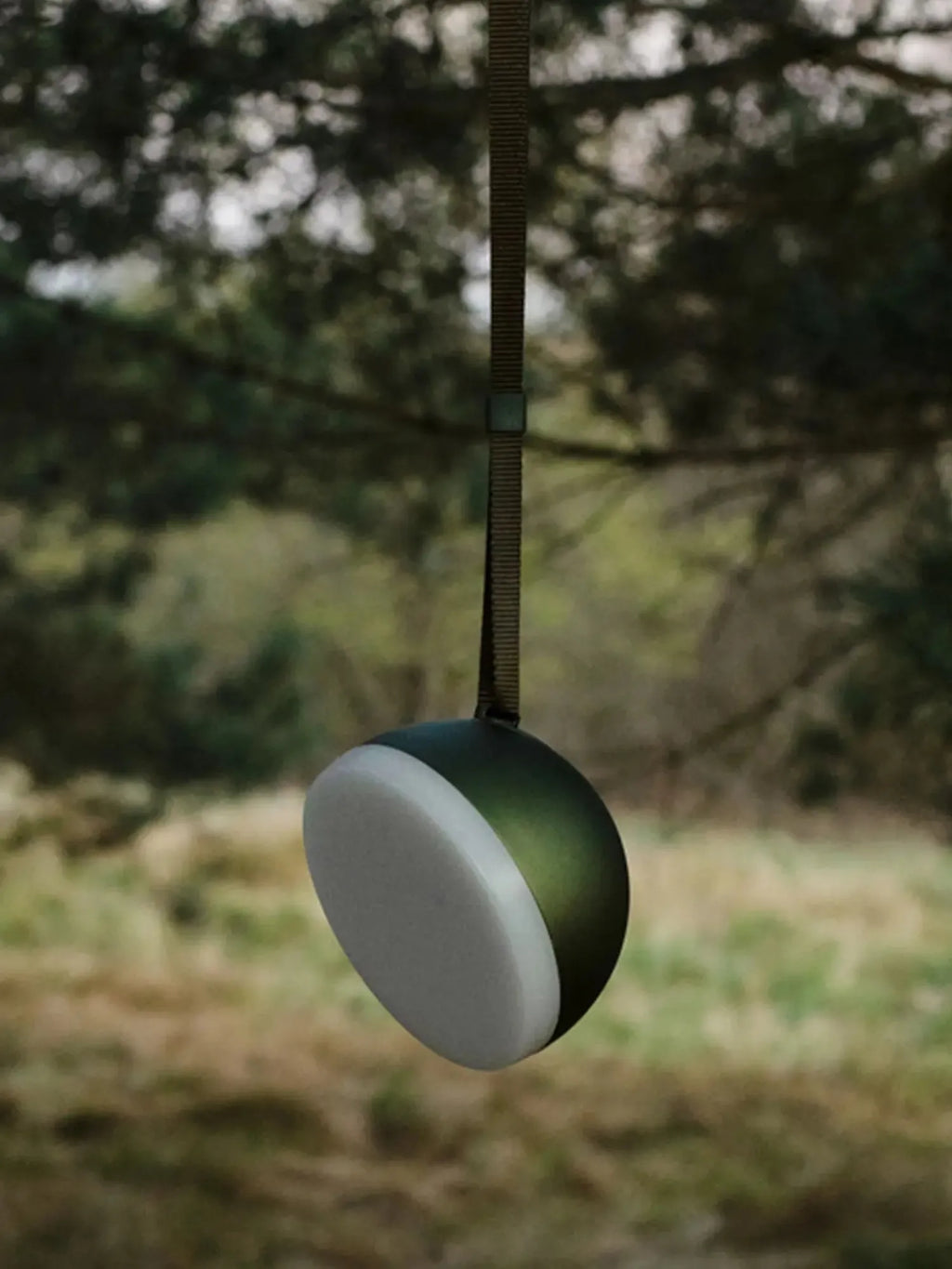 New Works Sphere Adventure Light, a premium dark green spherical lantern with a white translucent diffuser, hanging from a rugged dark strap against a blurred outdoor landscape. Ideal for outdoor adventures.