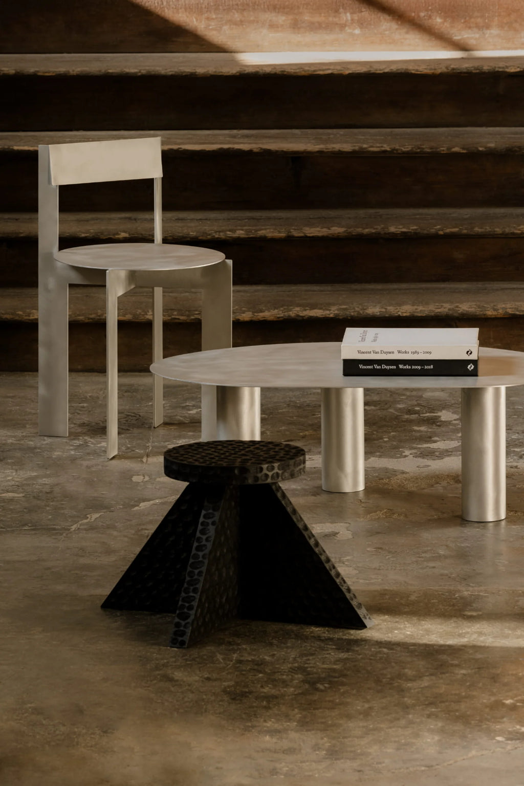NG DESIGN Object 112 Side Table, a premium matte silver circular metal table on stout cylindrical legs, styled with two books, next to a matching metal chair and a textured black sculptural stool on a concrete floor. Ideal for modern living spaces.