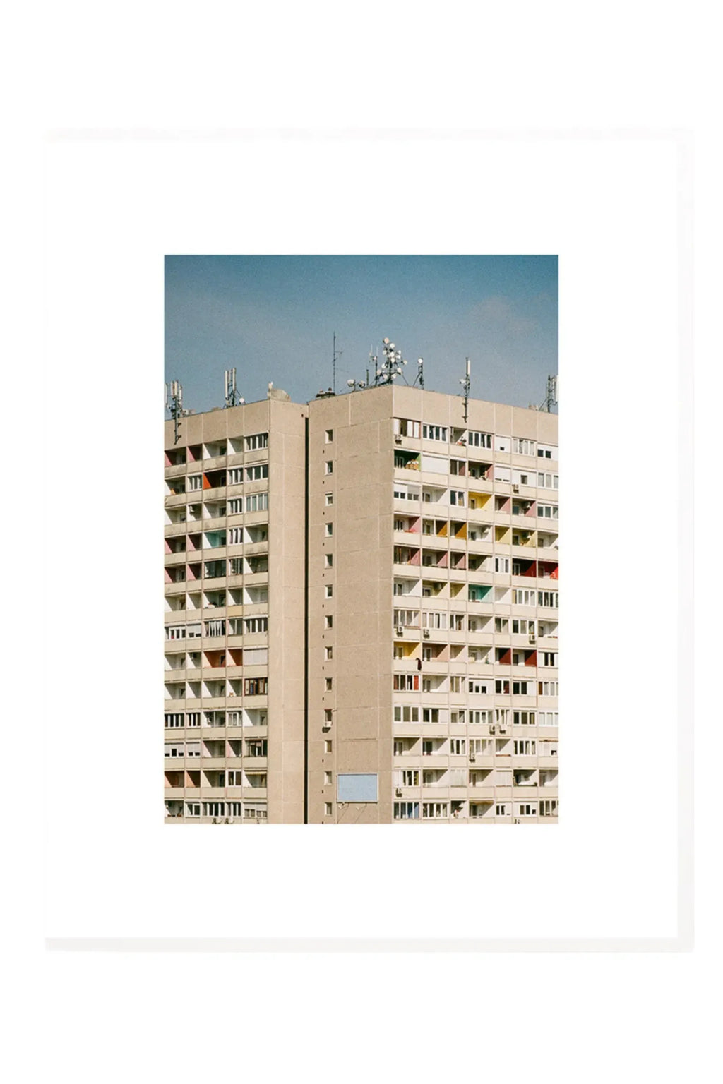 The Poster Club Block O’Clock, an architectural photography poster featuring a tall, brutalist concrete building with a beige facade under a clear blue sky. The building's numerous balconies and windows reveal vibrant pops of color in red, yellow, green, and blue, adding a contemporary touch to the urban landscape.