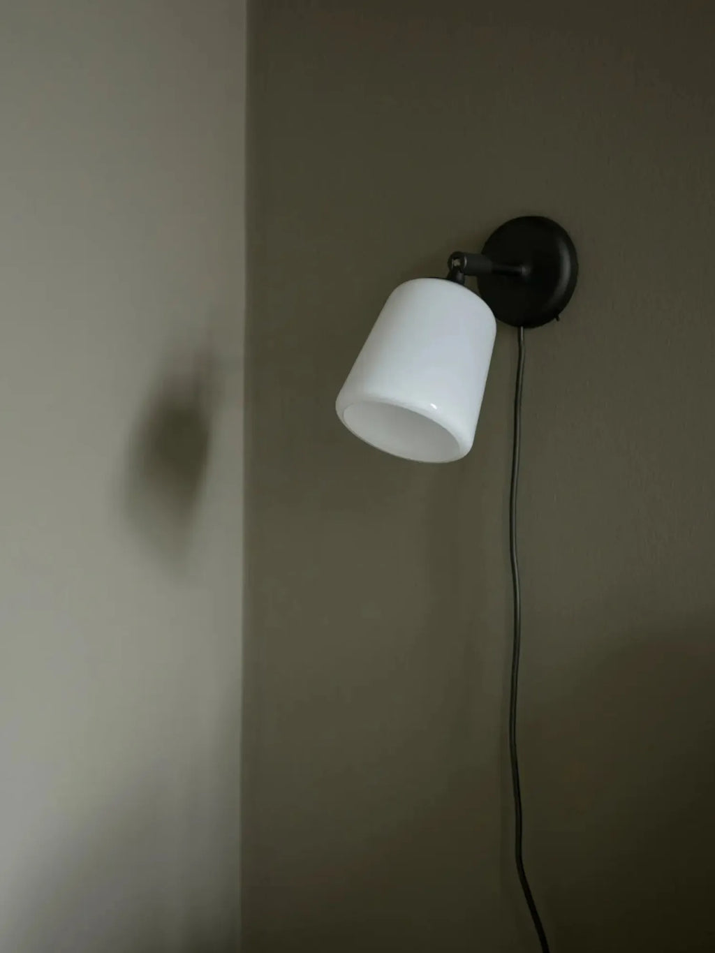 New Works Material Wall Lamp: Modern black metal base with an opaque white glass conical shade, featuring a visible black cable, mounted on a dark olive green wall. Provides premium adjustable lighting.
