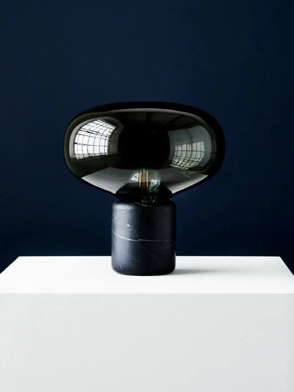 New Works Karl-Johan Table Lamp, showcasing its smoked black glass shade and dark marble base with white veining, displayed on a clean white surface. A striking, luxurious design for modern interiors.