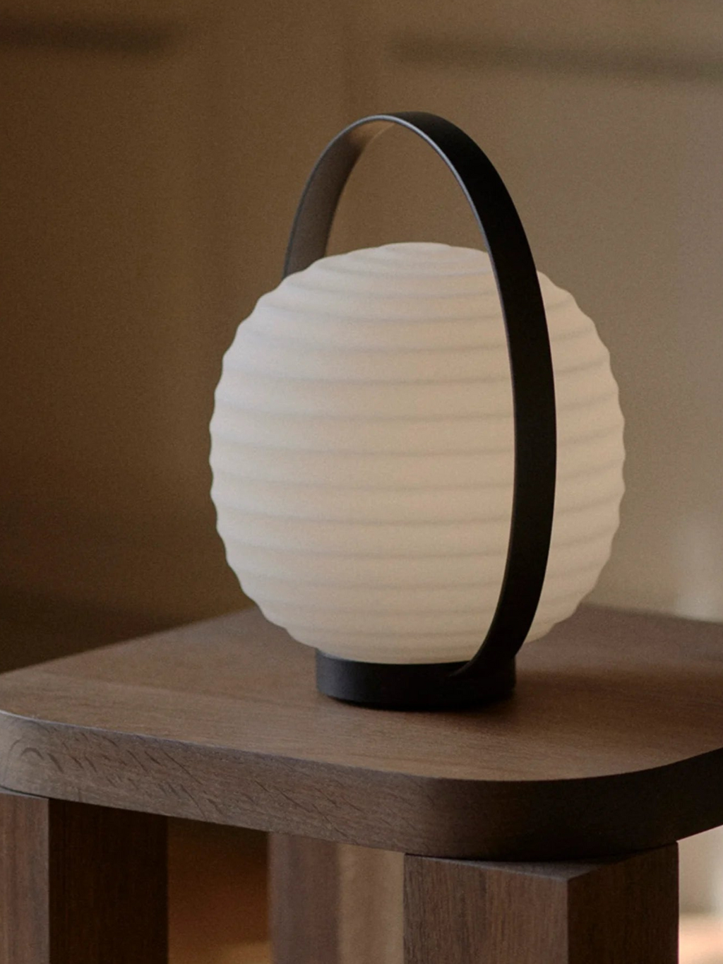 New Works Lantern Portable Lamp with a spherical, ribbed opaque white glass diffuser and matt black metal handle, displayed on a wooden side table. This premium battery-powered lamp offers ambient, versatile lighting.
