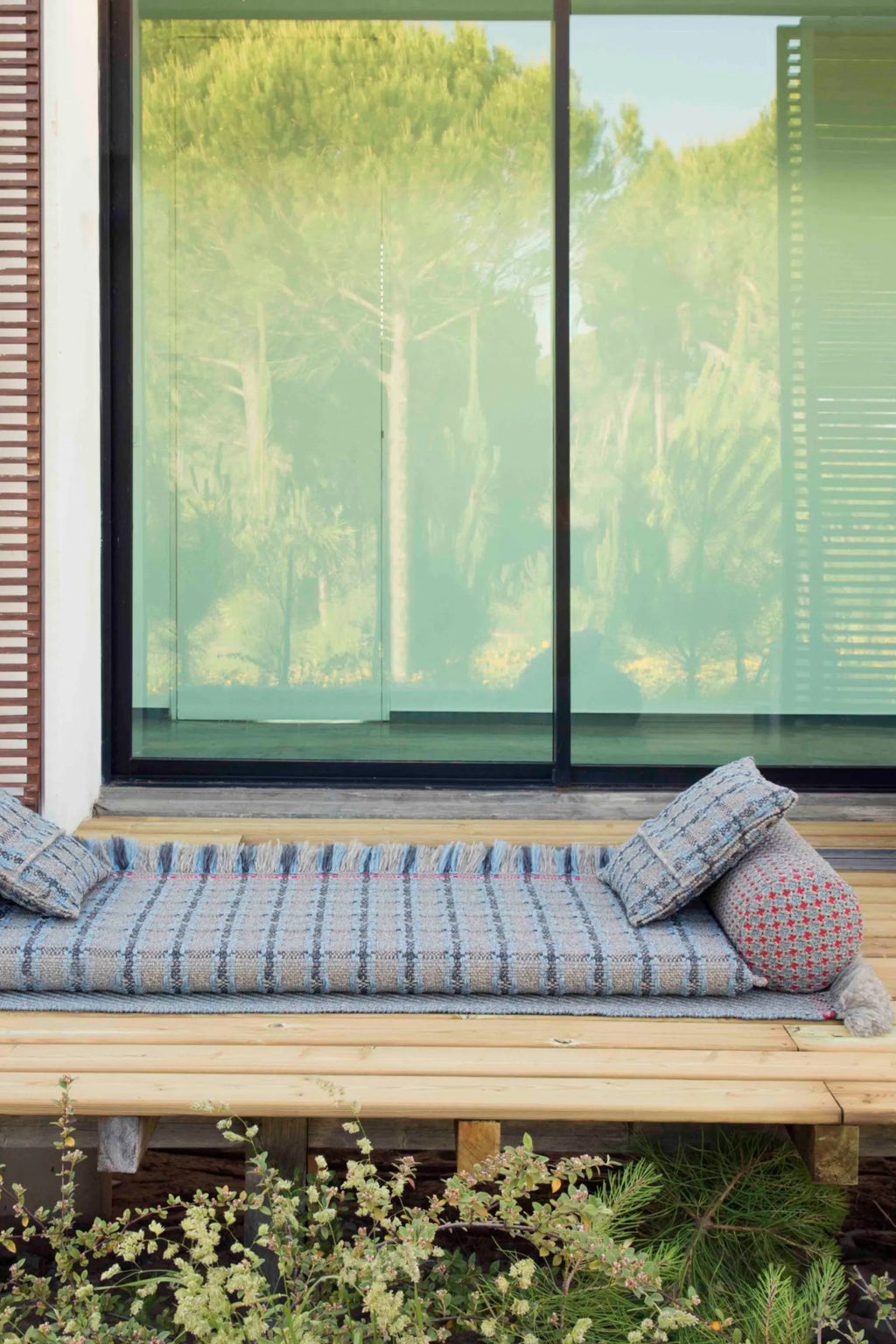 GAN Rugs Garden Layers Composition 05, a contemporary outdoor sofa featuring a hand-woven grey and blue striped mattress with fringed edges, crafted from durable outdoor fabric with an organic texture. It includes a rectangular throw pillow and a cylindrical bolster pillow, both in coordinating patterns with red accents. Perfect as an outdoor daybed or lounge cushion for a rustic chic patio, balcony, or garden living space.