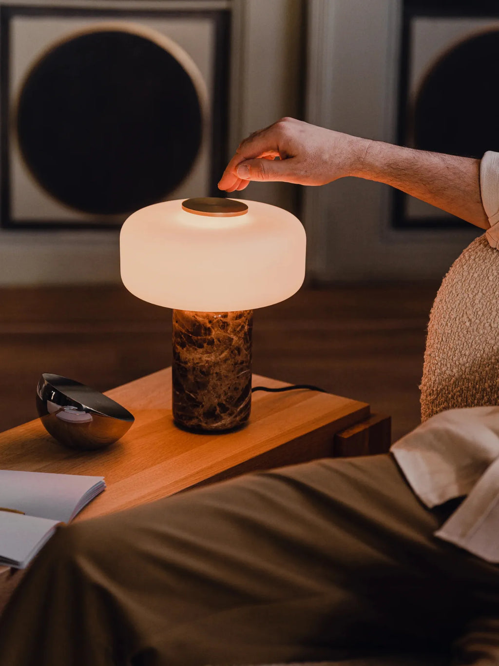 TAMO Fonte Table Lamp, an elegant modern lamp with a glowing white mushroom-shaped shade & a luxurious dark brown marble base, being touched by a hand. A perfect luxury decor piece.
