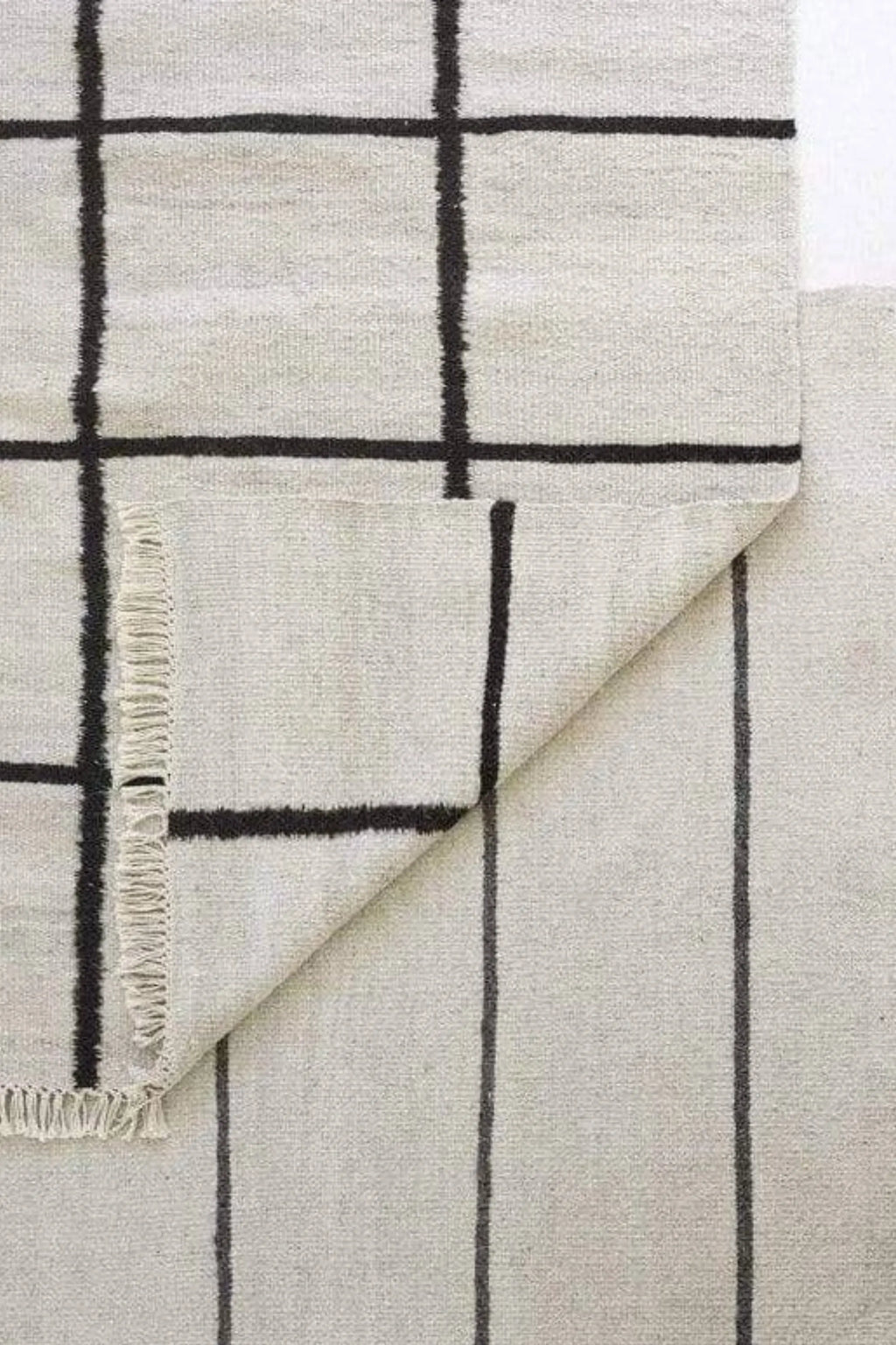 Sera Helsinki Kuura Rug, a modern minimalist hand-woven flatweave rug crafted from natural wool in a light grey hue, featuring a distinctive dark charcoal grid pattern and subtle fringe details.