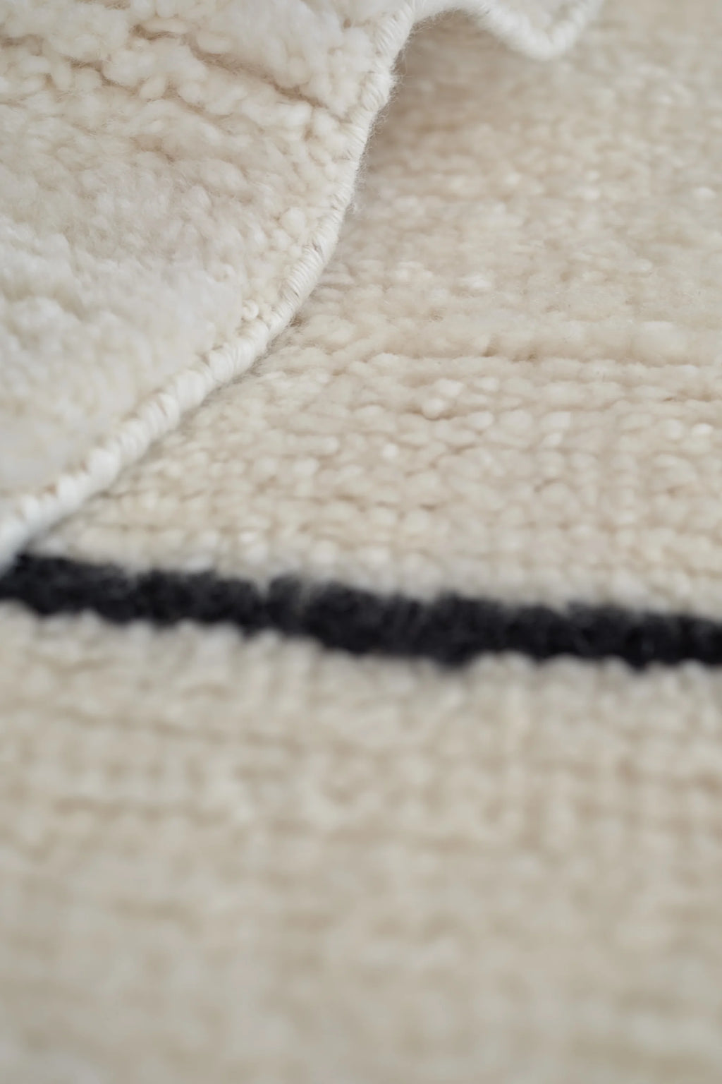 Linie Design Astral Spiral Wool Rug, a plush, hand-tufted wool area rug in a soft natural cream color with a distinctive bold black stripe. It showcases a thick, shaggy pile and an inviting, cozy texture, ideal for adding warmth and a modern Scandinavian aesthetic to any space. Perfect as a minimalist bedroom rug, a contemporary living room accent, or for creating a Nordic-inspired interior.