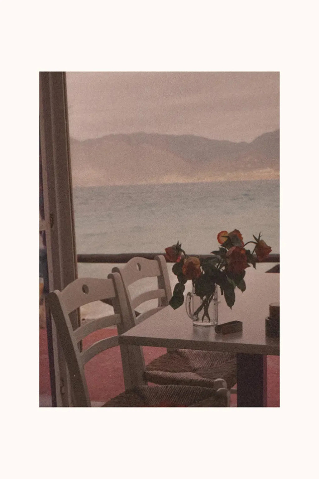 The Poster Club Dine with Me, a nostalgic art print featuring a vintage film photography scene of a coastal cafe table with light wood chairs and a clear glass vase of faded red roses, overlooking a serene blue sea and distant mountains under a soft, muted pink and grey sky.