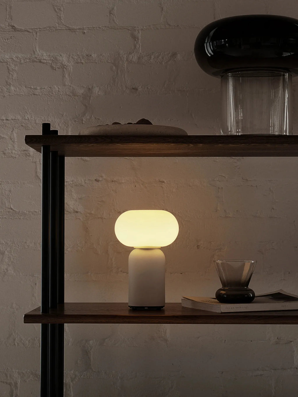 TAMO Velis Portable Lamp, a modern, glowing orb with a beige ceramic base, casting a warm light on a dark wooden shelf against a white brick wall. Perfect for creating ambient lighting.