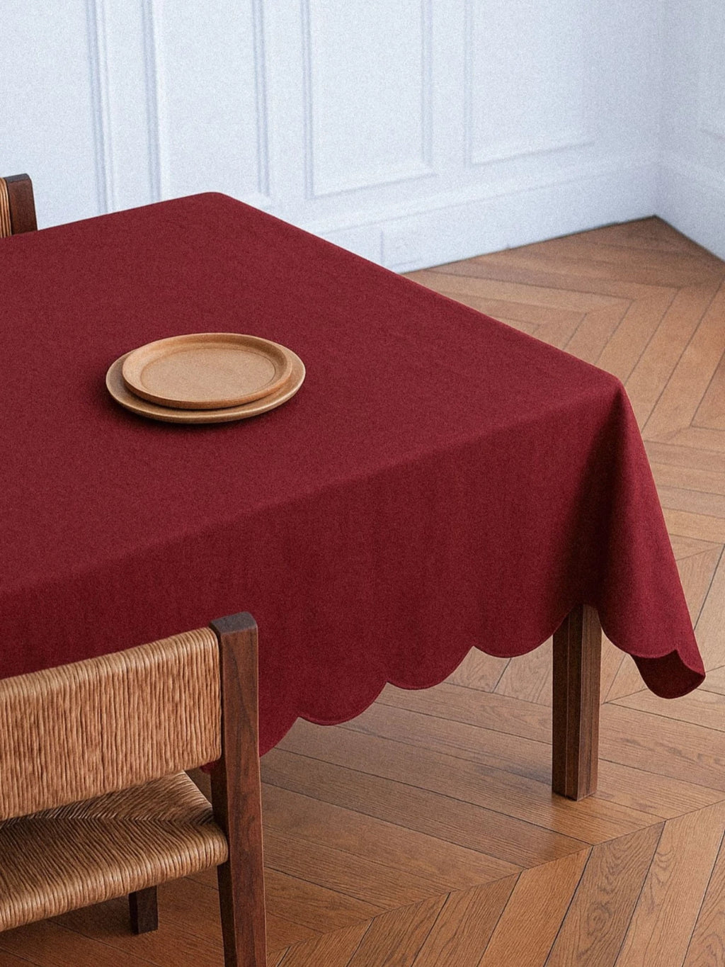A square dining table is covered with a deep burgundy or maroon Scalloped Linen Tablecloth that features a decorative wavy, scalloped edge. Two simple, light-brown wooden plates are stacked in the upper left corner of the tablecloth. A dark wood chair with a woven rush seat and back is visible in the foreground. The floor is made of warm, light-brown wood parquet, and the background wall is white with subtle decorative molding.