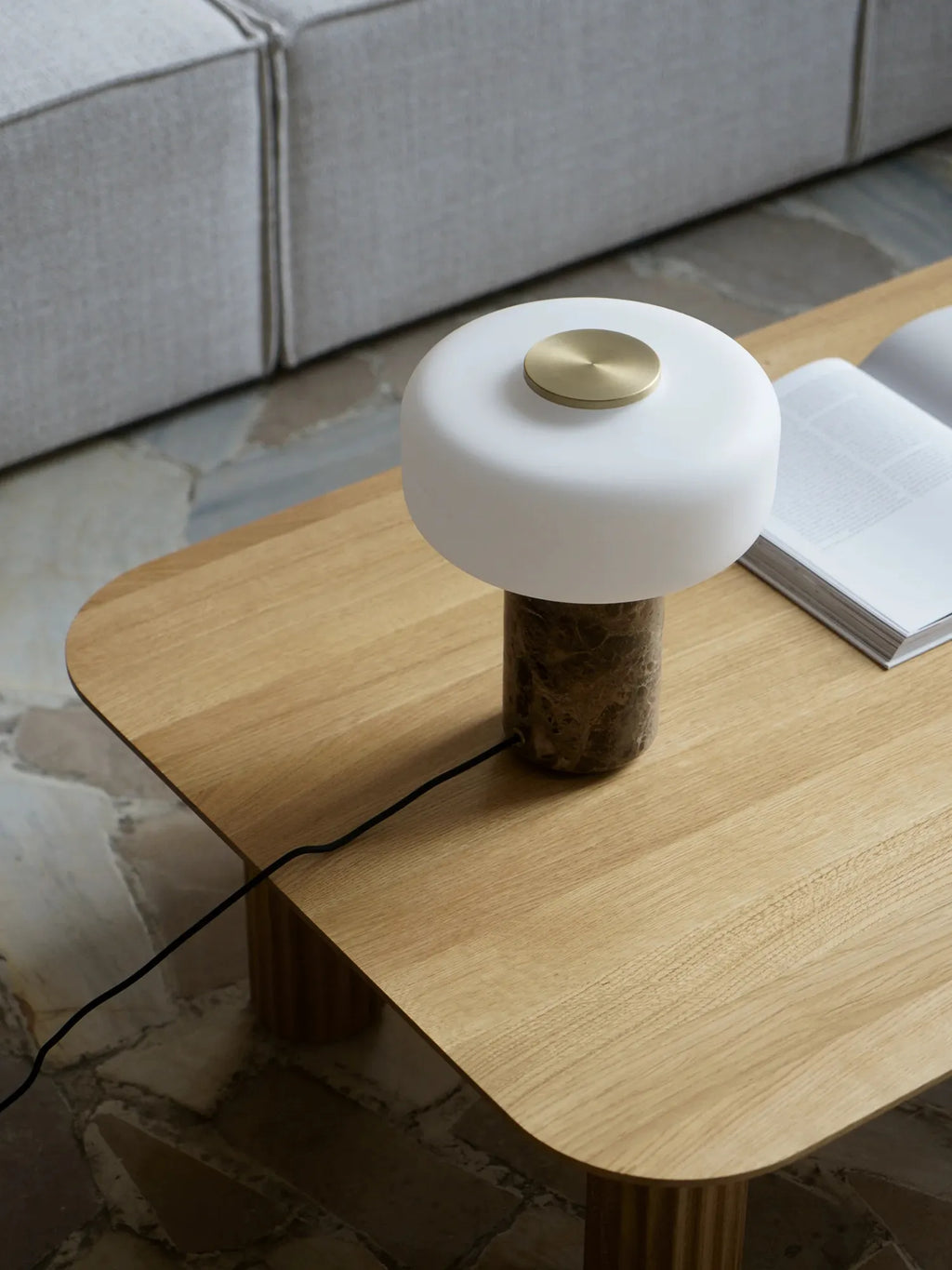 TAMO Fonte Table Lamp, a luxurious modern lamp with a translucent white dome diffuser, solid brass top accent, and a dark brown marble base, displayed on a light wooden coffee table. Ideal for ambient lighting.