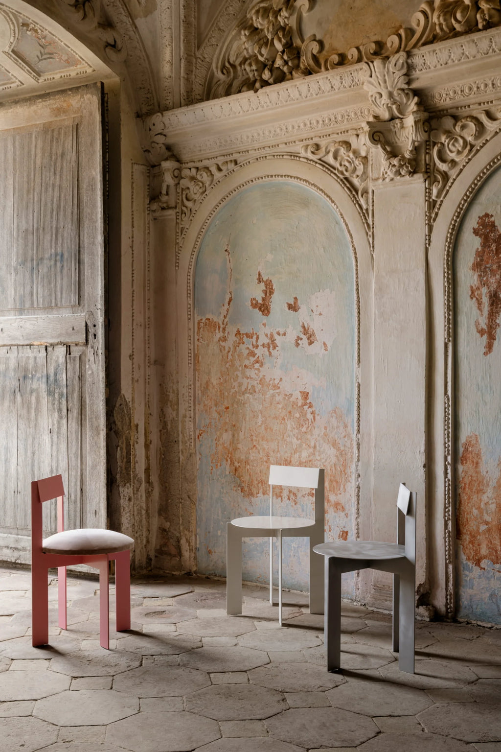 NG DESIGN Object 111 Chair set: a pink chair with plush round seat cushion, a white metal chair, and a grey minimalist chair, showcased on an antique stone floor in a rustic, distressed room. Premium modern seating.