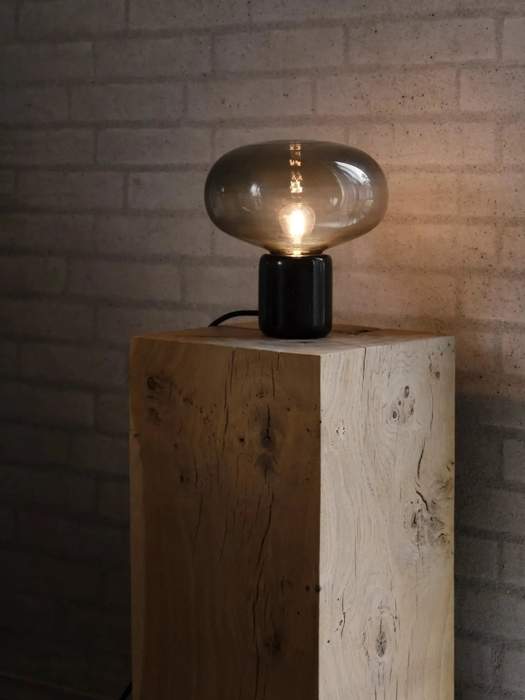 New Works Karl-Johan Table Lamp with a smoked glass bulb on a matte black base, sitting on a rustic light wood block. This luxury lamp offers ambient lighting.
