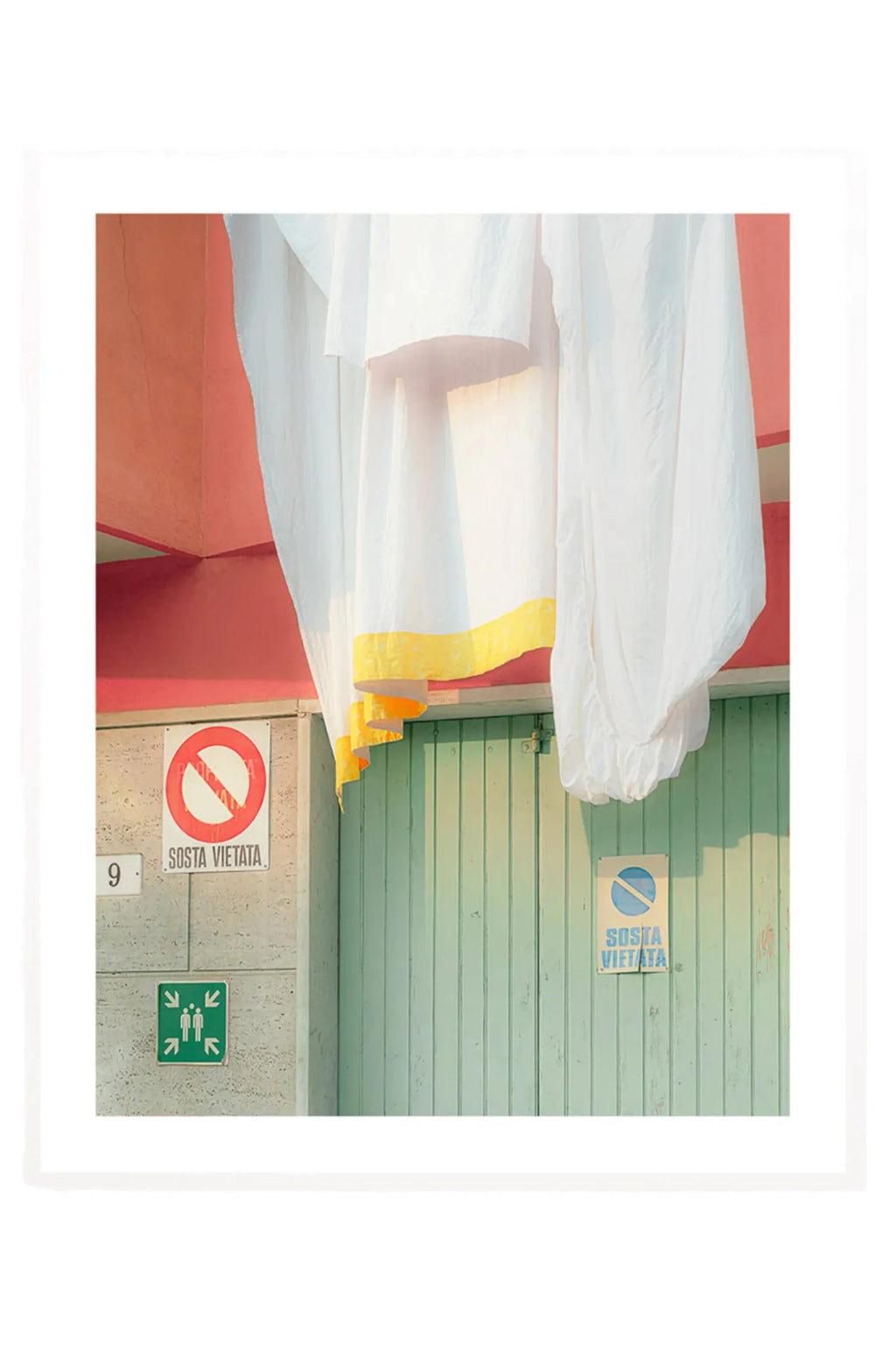 The Poster Club Camogli - Photo Art Print, a vibrant coastal photography print capturing a sun-drenched Italian street scene with a textured coral-red wall, a weathered mint green wooden door, and flowing white laundry featuring a bright yellow border. This Mediterranean art print offers a minimalist composition with striking color blocking, perfect for gallery wall art or as a vibrant accent in coastal or contemporary interiors.