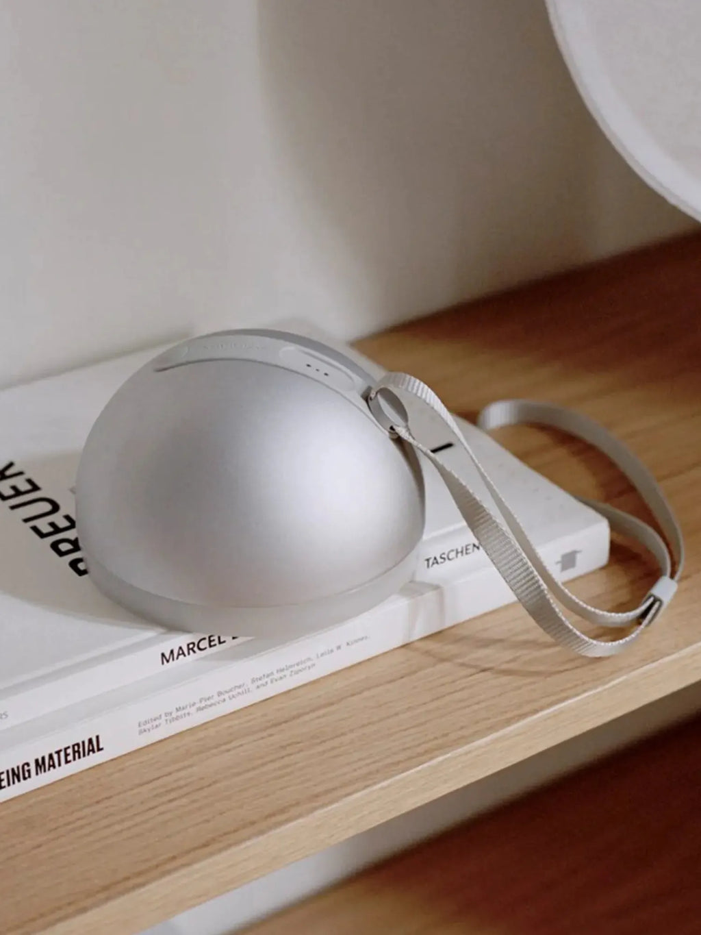 New Works Sphere Adventure Light, a sleek silver spherical portable lamp with a subtle textured base and a light gray woven strap, shown styled on white books on a wooden shelf. A premium, modern lighting solution.