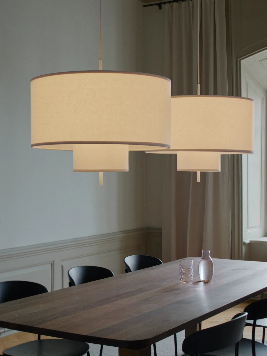 New Works Margin Pendant Lamp, featuring two layered round drum shades in natural white fabric with subtle brown trim, suspended over a dark wood dining table. Provides elegant, ambient light.