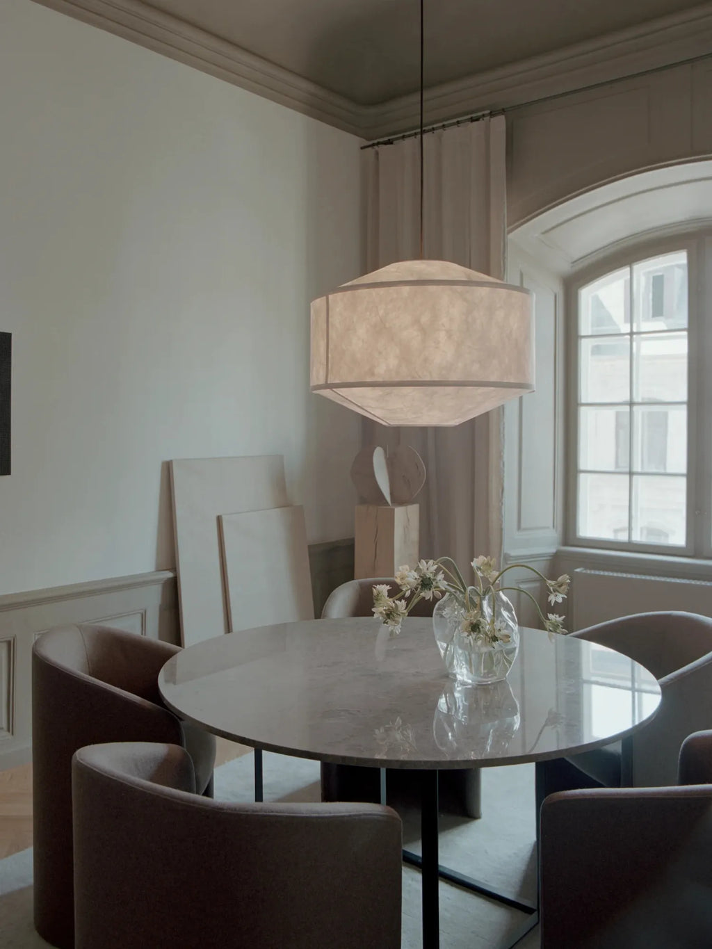 New Works Kite Pendant Lamp; a large, trapezoidal ceiling-mounted light with a textured, translucent white paper shade, suspended over a modern dining table. This luxury lighting piece offers ambient illumination.