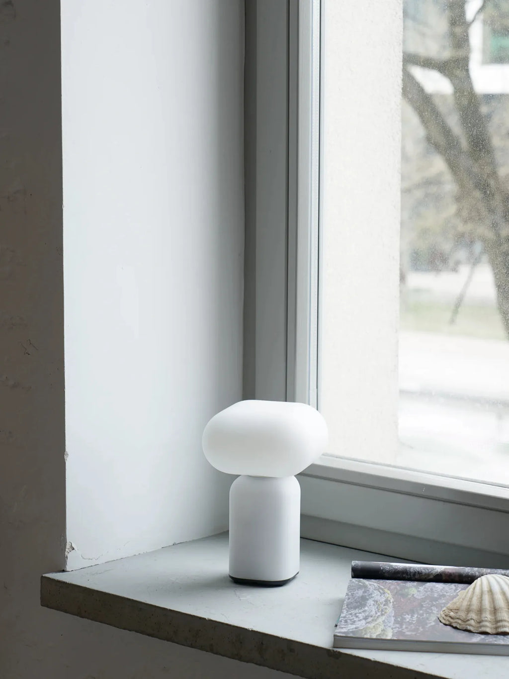 TAMO Velis Portable Lamp, a minimalist modern white mushroom-shaped light with a matte finish, shown on a concrete windowsill next to a magazine and a seashell. Offers warm, ambient lighting.
