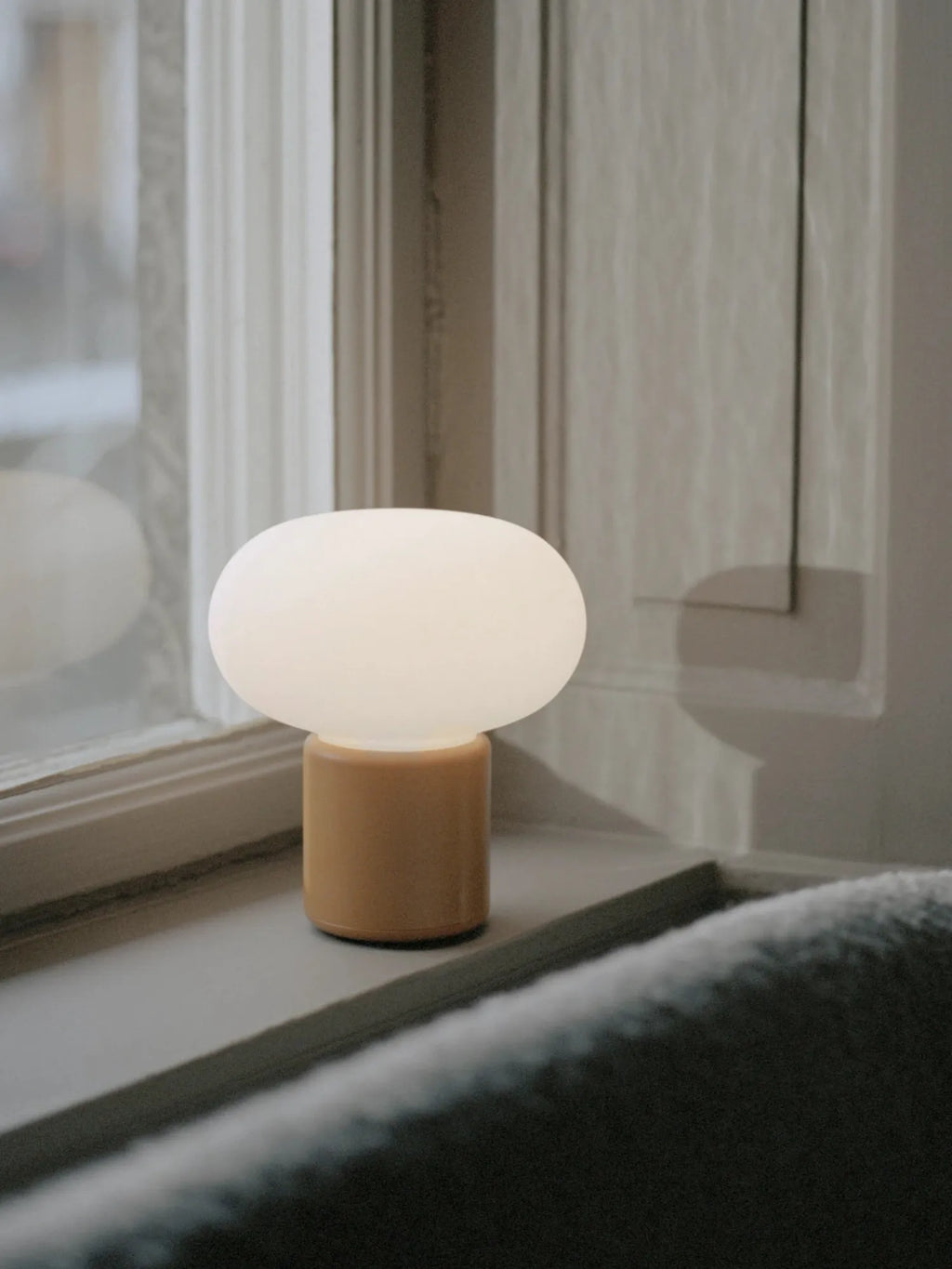 New Works' Karl-Johan Portable Table Lamp with a milky white glass shade and an amber cylindrical base, displayed on a windowsill. This premium modern lamp is ideal for ambient lighting.