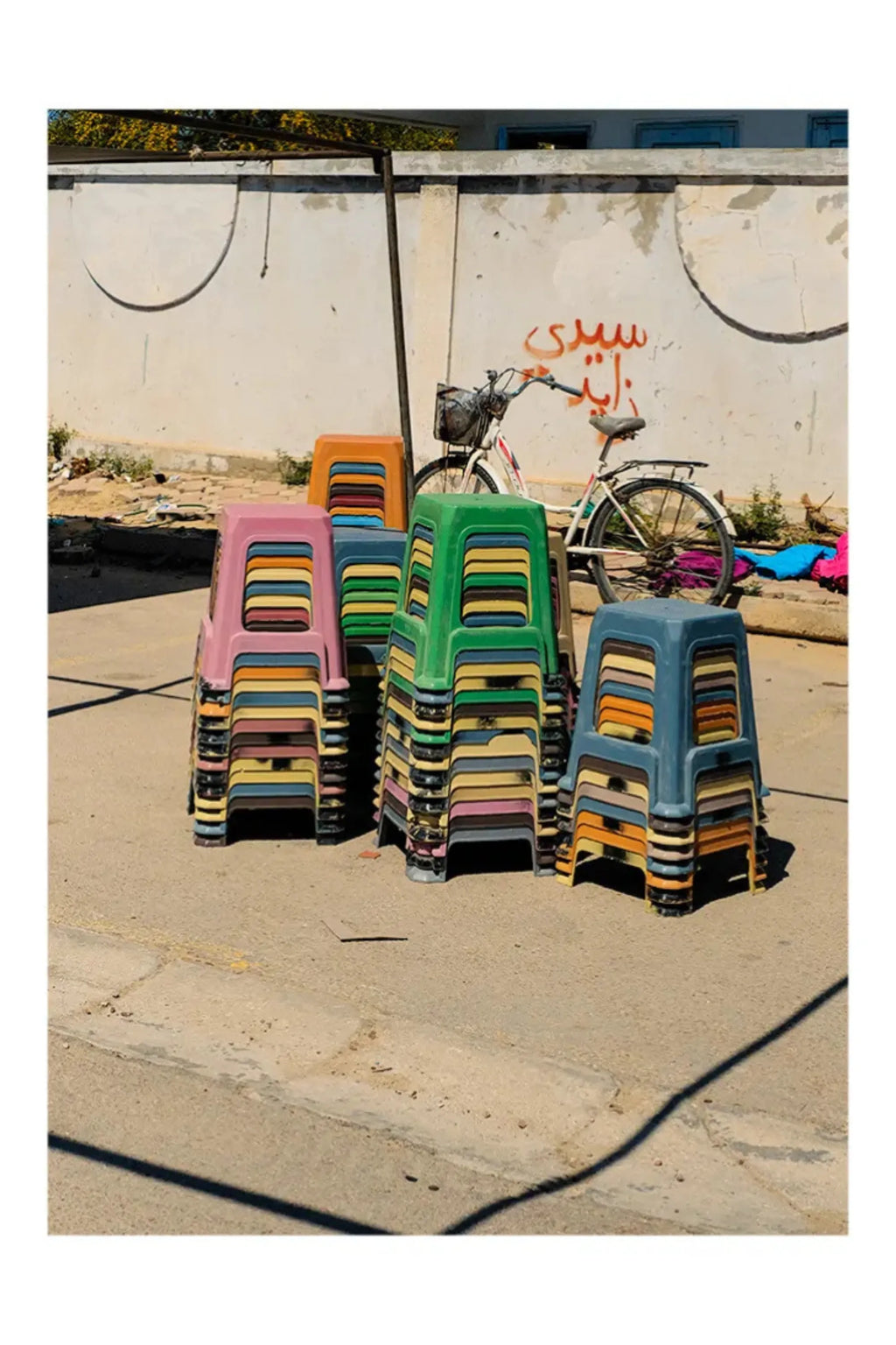The Poster Club The Souk, a vibrant street photography poster capturing a bustling market scene with towering stacks of colorful plastic stools in shades of pink, green, blue, orange, and yellow.